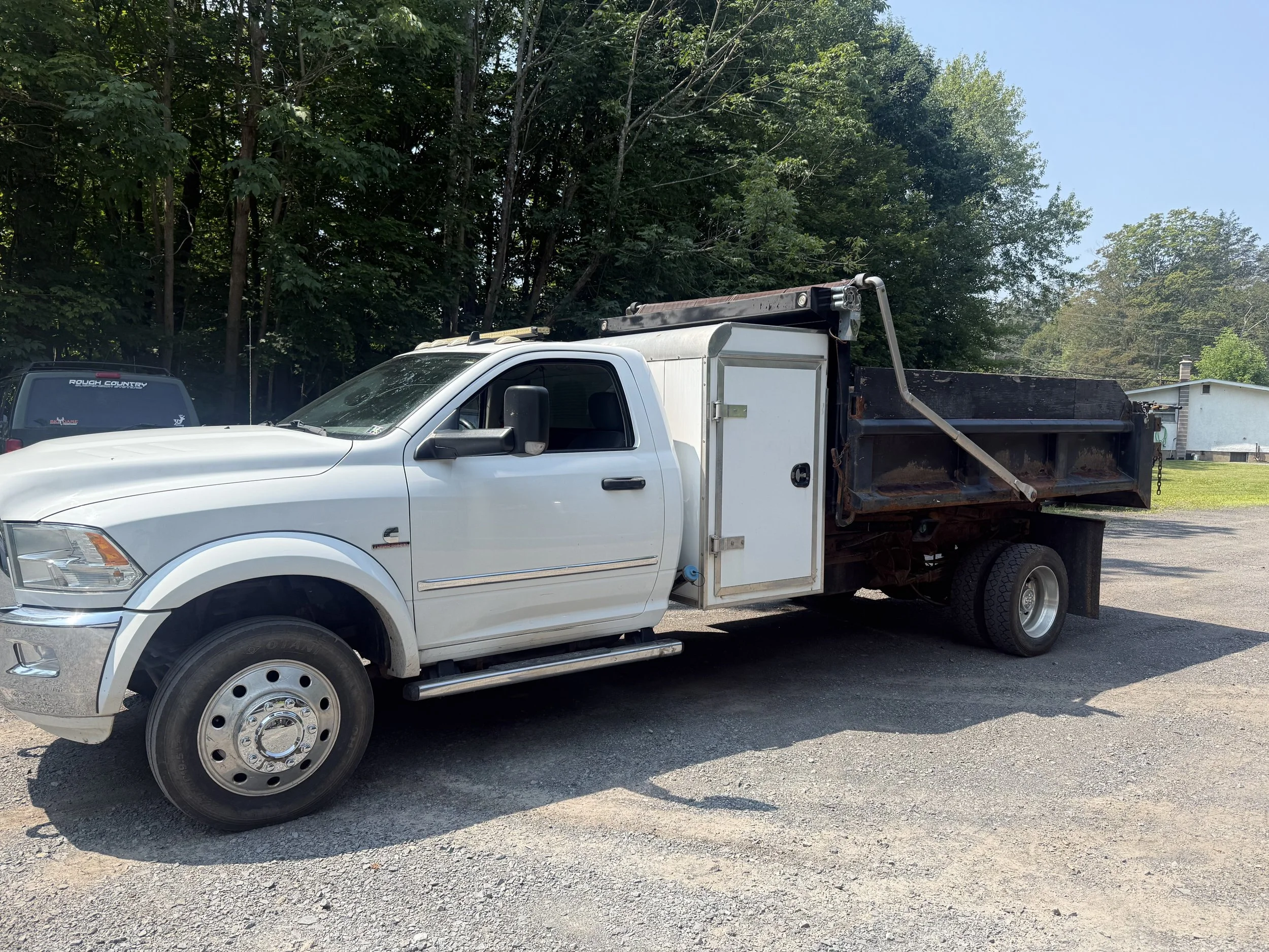 A white flatbed truck with a black bed and side storage compartment parked on a gravel surface with trees and a building in the background.