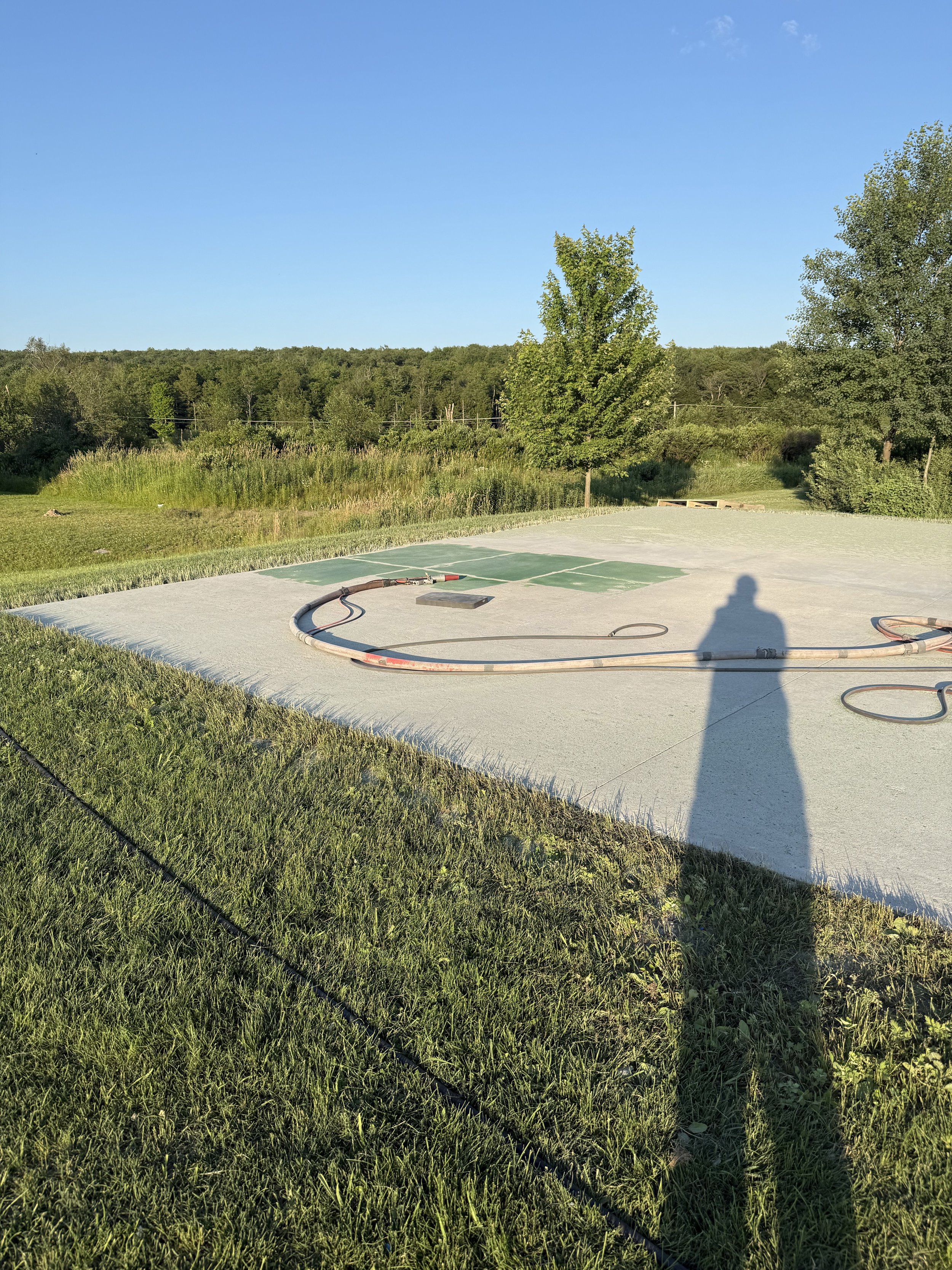 A sports court under construction with some equipment on it, surrounded by grass and trees on a sunny day.