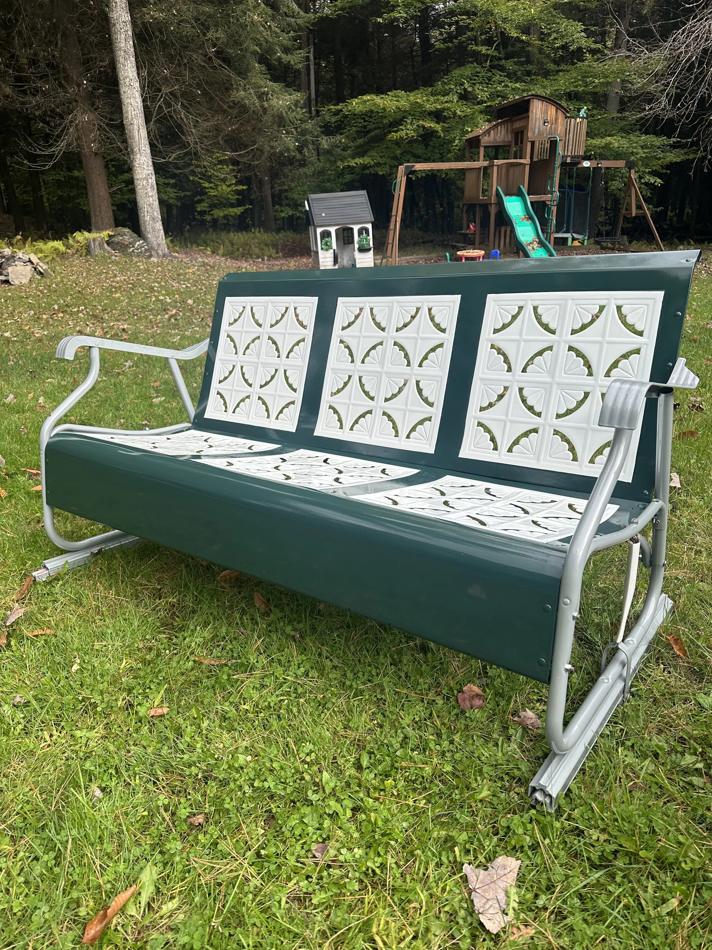 A metal and plastic outdoor bench with a dark green backrest and white decorative panels on the seat and back, placed on a grassy lawn with a backyard playset and trees in the background.