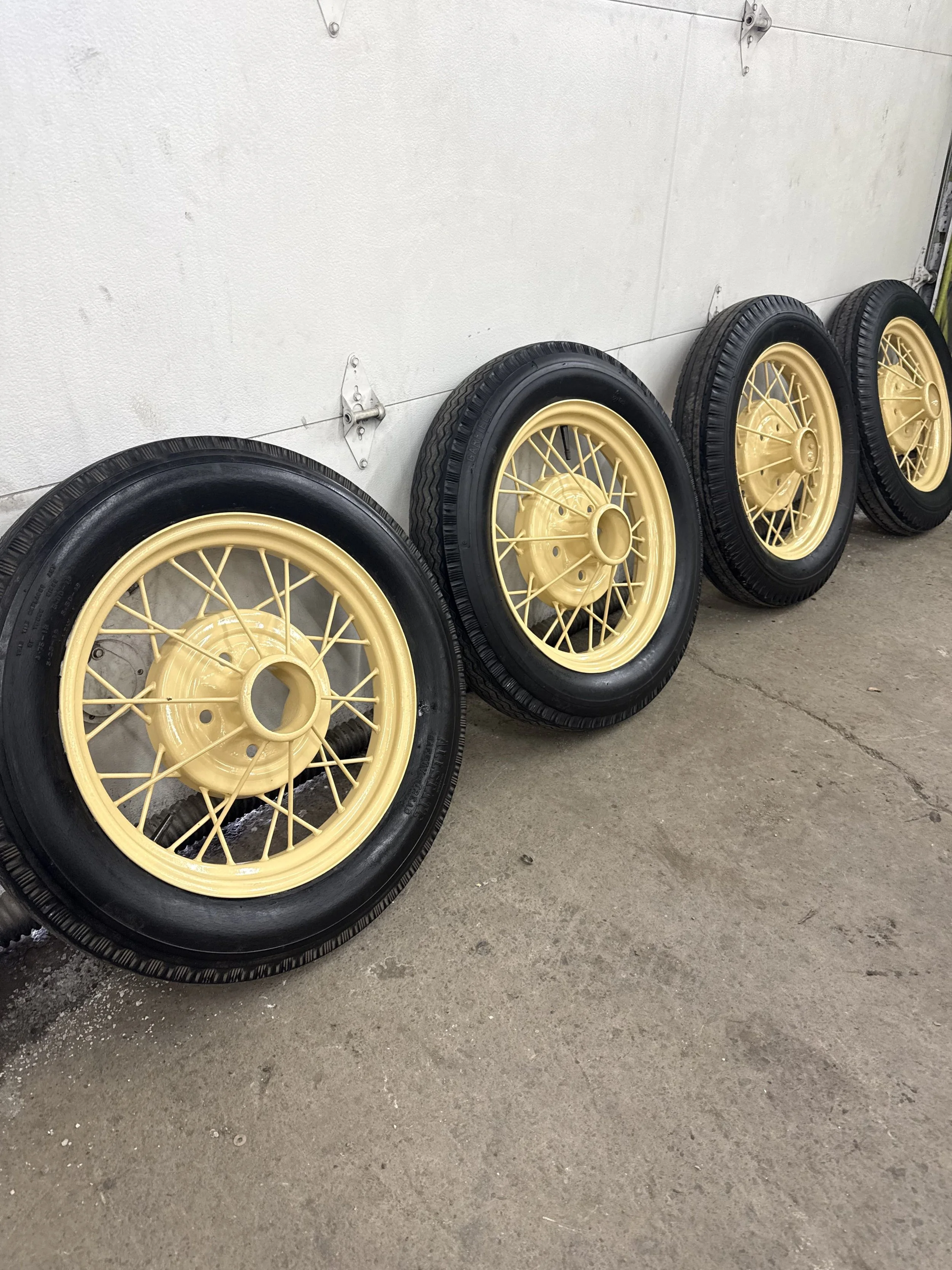 Four vintage-style car wheels with black tires and yellow wire spokes, lined up against a white wall in a garage or workshop.