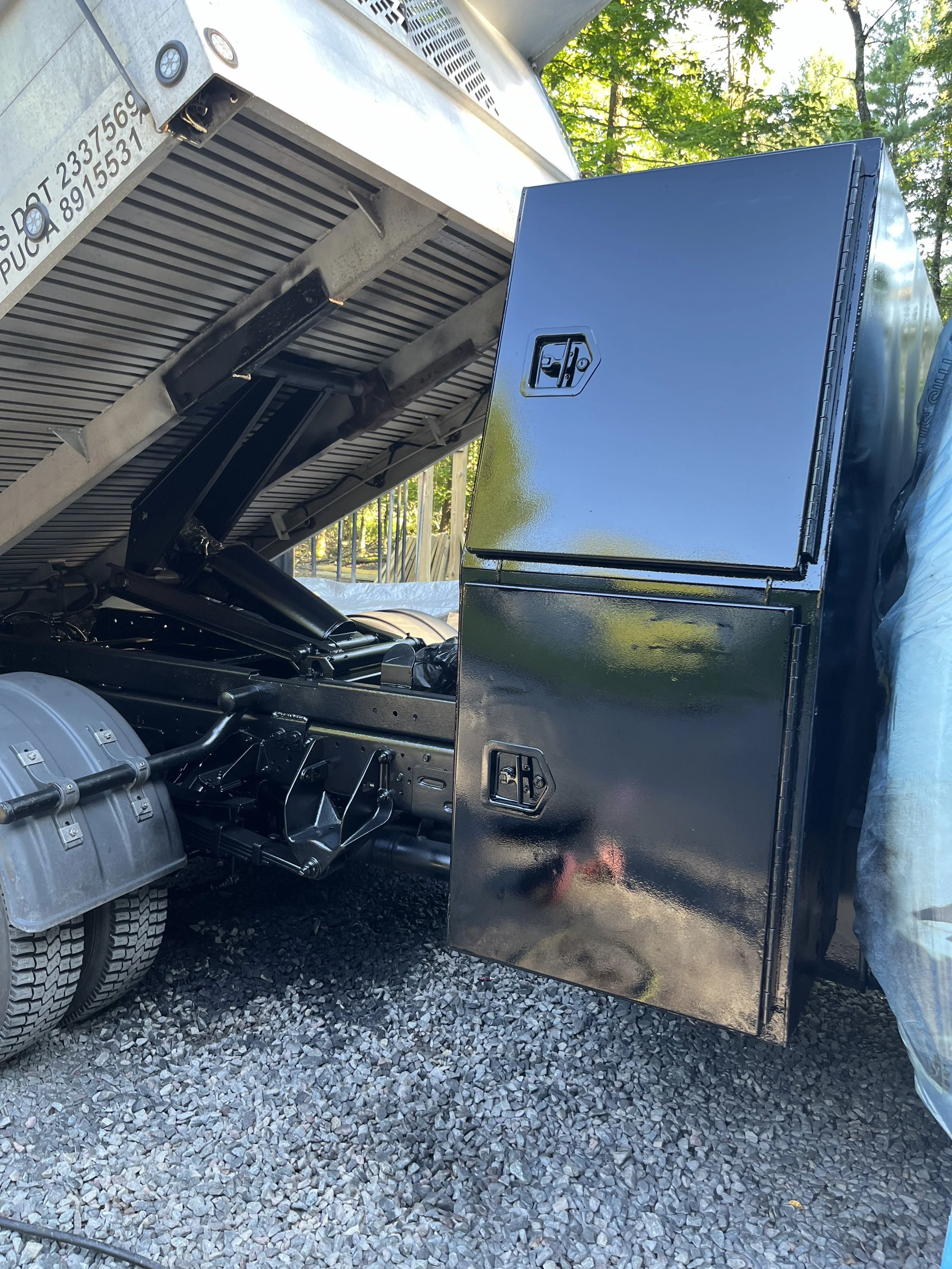 Close-up of a black toolbox attached to the side of a silver flatbed truck, with hydraulic components and gravel ground visible.