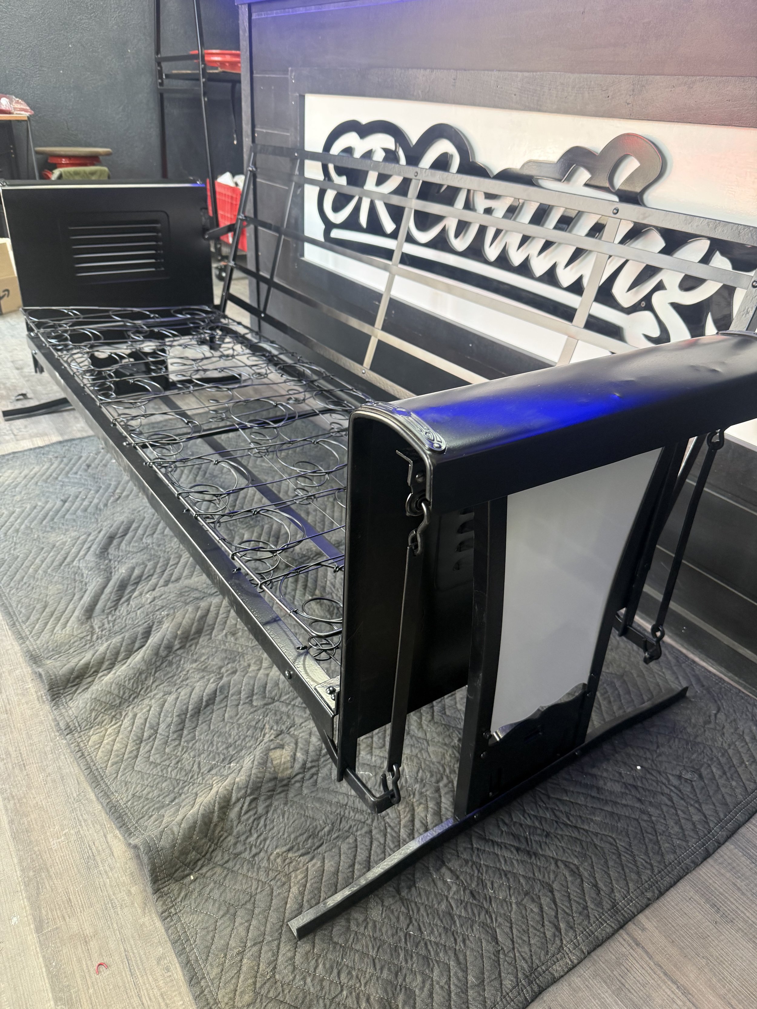 Frame of a black metal bed with a wire mesh base, positioned on a grey padded quilt on the floor.