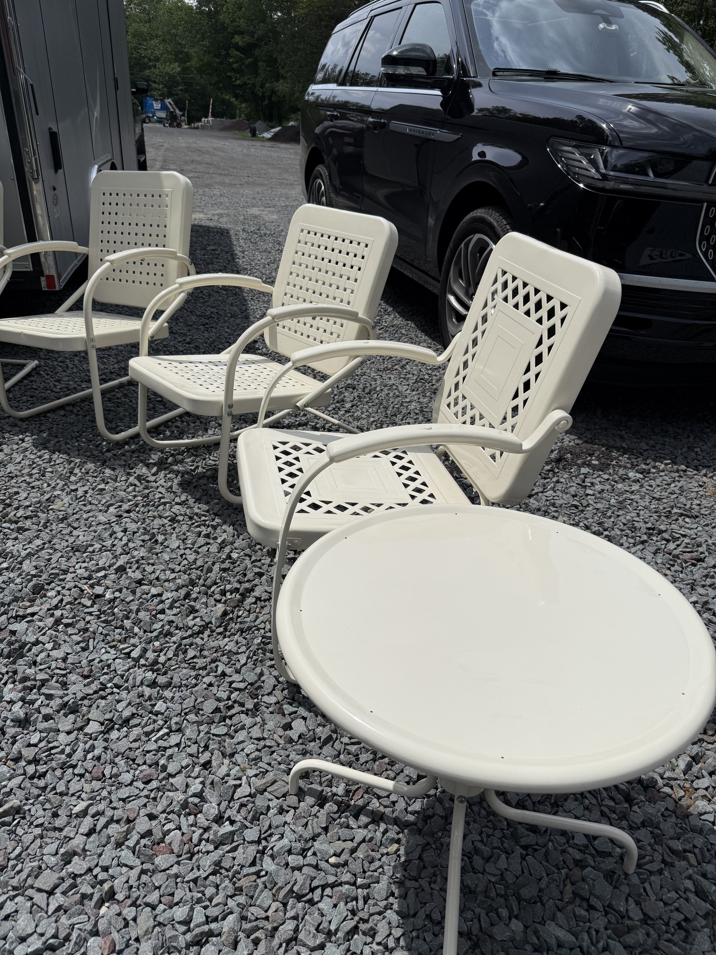 A set of white outdoor patio furniture including four chairs and a round table on a gravel surface next to a black SUV vehicle.