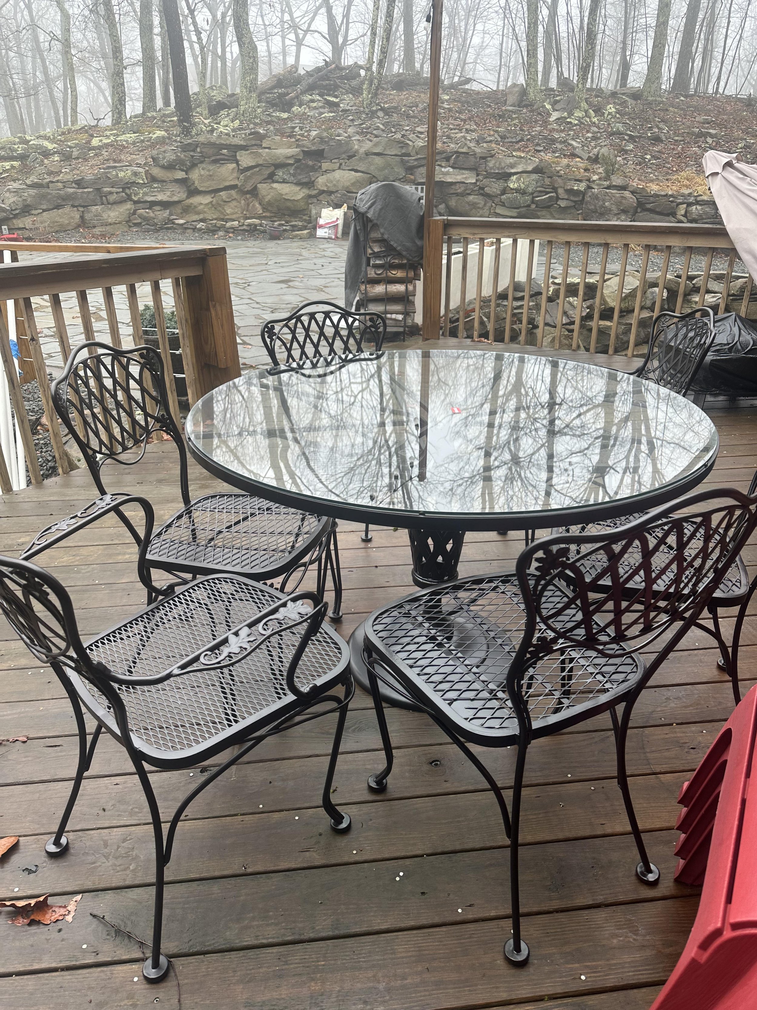 A wooden deck with a glass-top round table and six black metal chairs. The background shows an outdoor stone patio, a low stone wall, and a foggy wooded area with leafless trees.