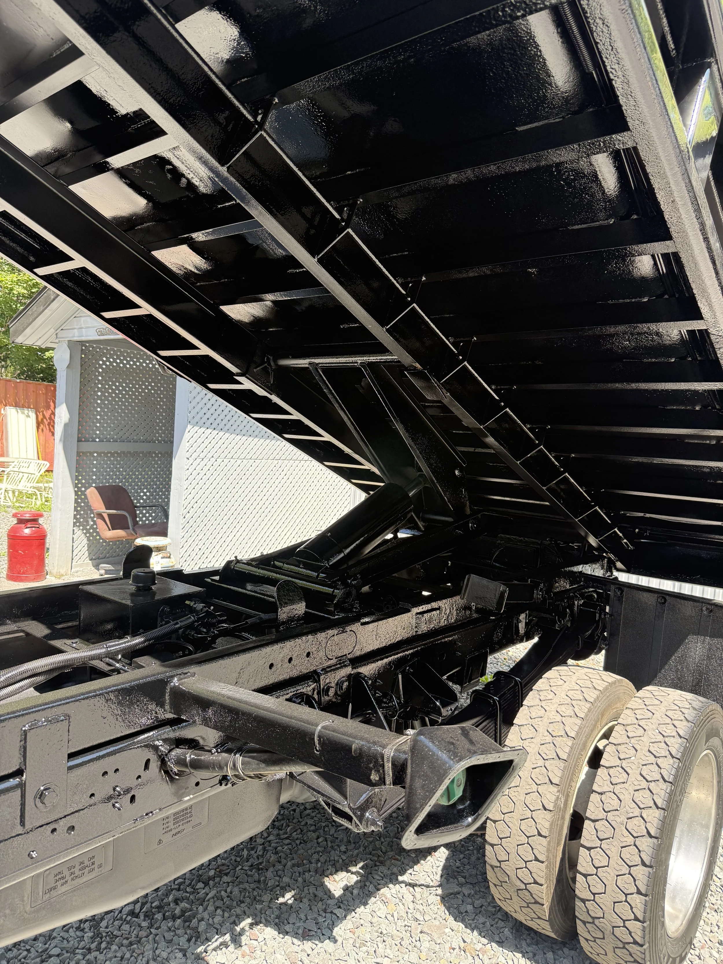 Close-up of a black dump truck bed tilted upward to show hydraulic lift mechanism and suspension system, with gravel ground and some outdoor furniture in background.