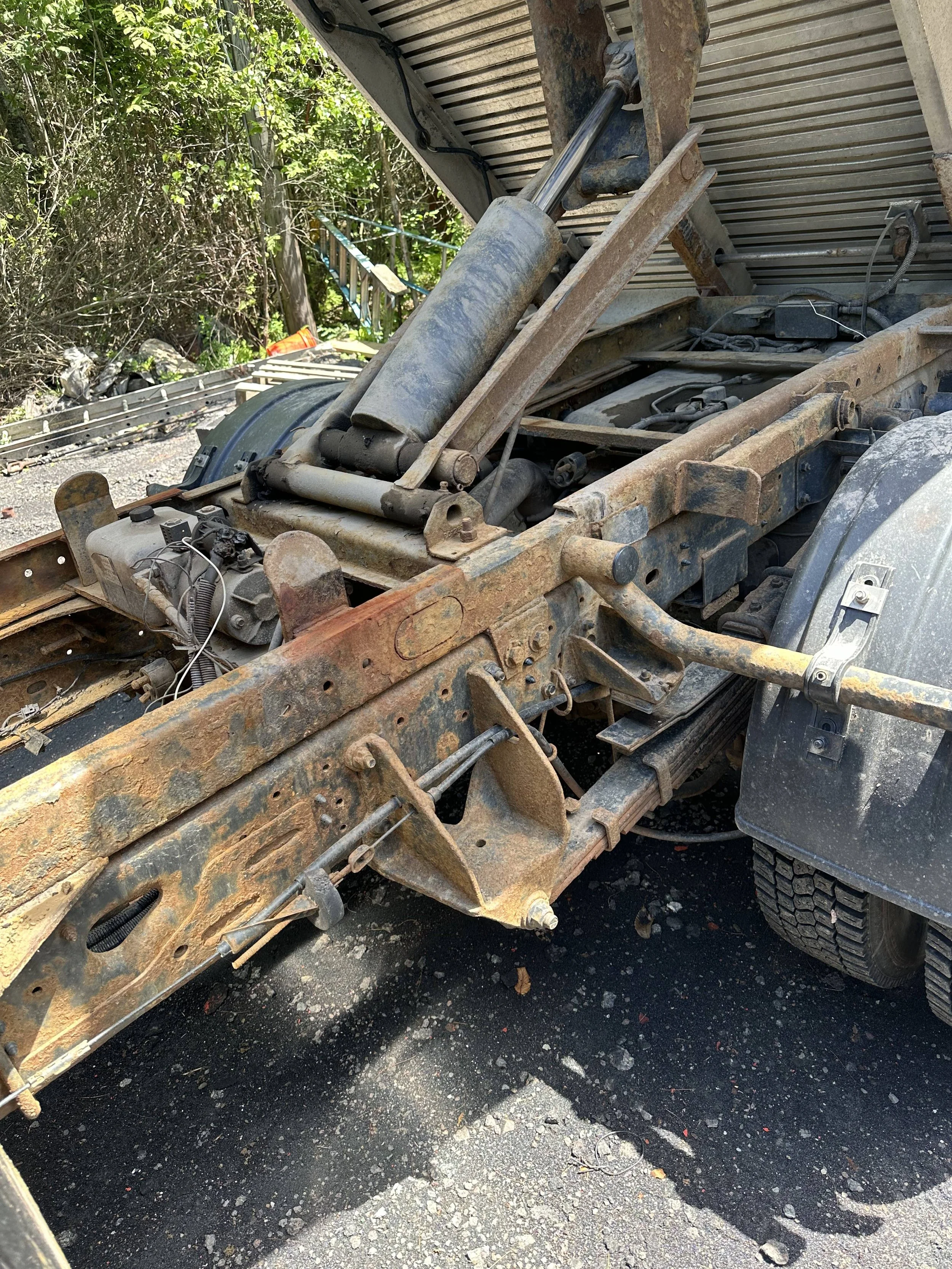 Rusty, partially dismantled truck chassis with visible suspension components, hydraulic cylinder, and frame, parked outdoors on asphalt with greenery and trees in the background.