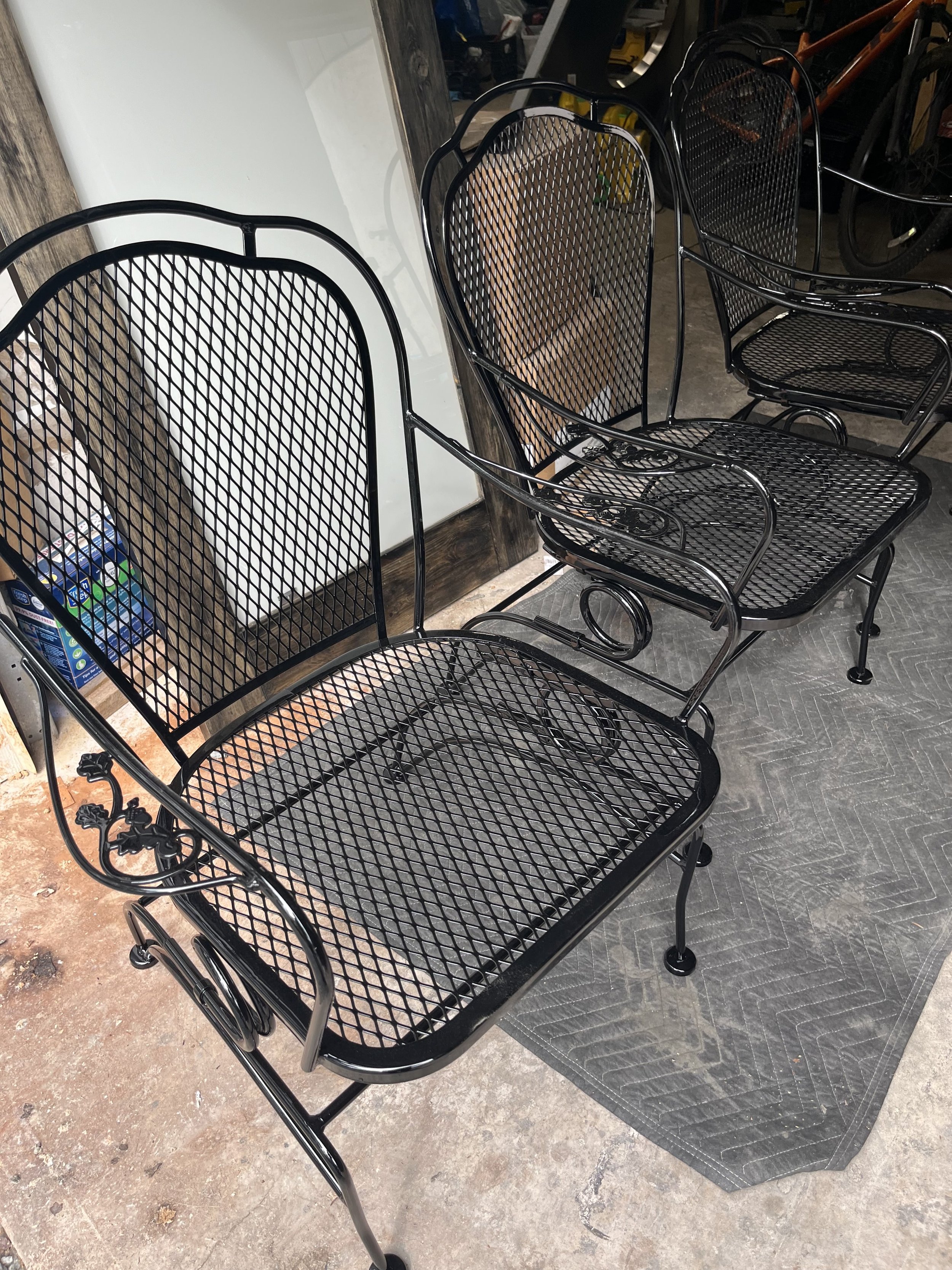 Black wrought iron patio chairs with mesh seats and backs, placed outdoors on a concrete floor next to a white wall and a wooden post.