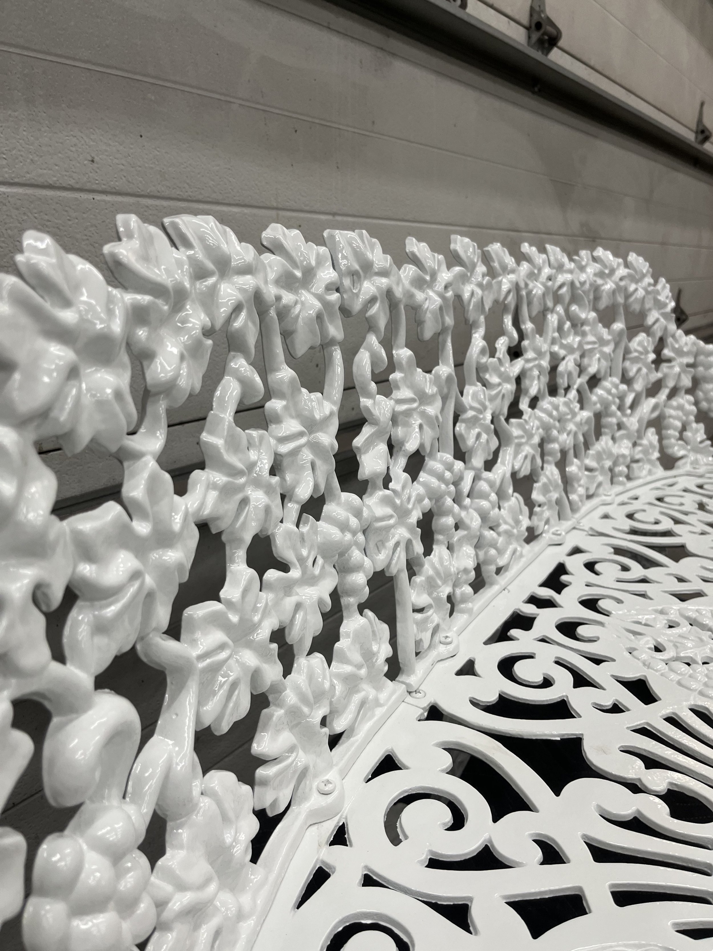 Close-up of a white ornate metal garden bench with decorative floral and scroll patterns, set against a gray wooden wall.
