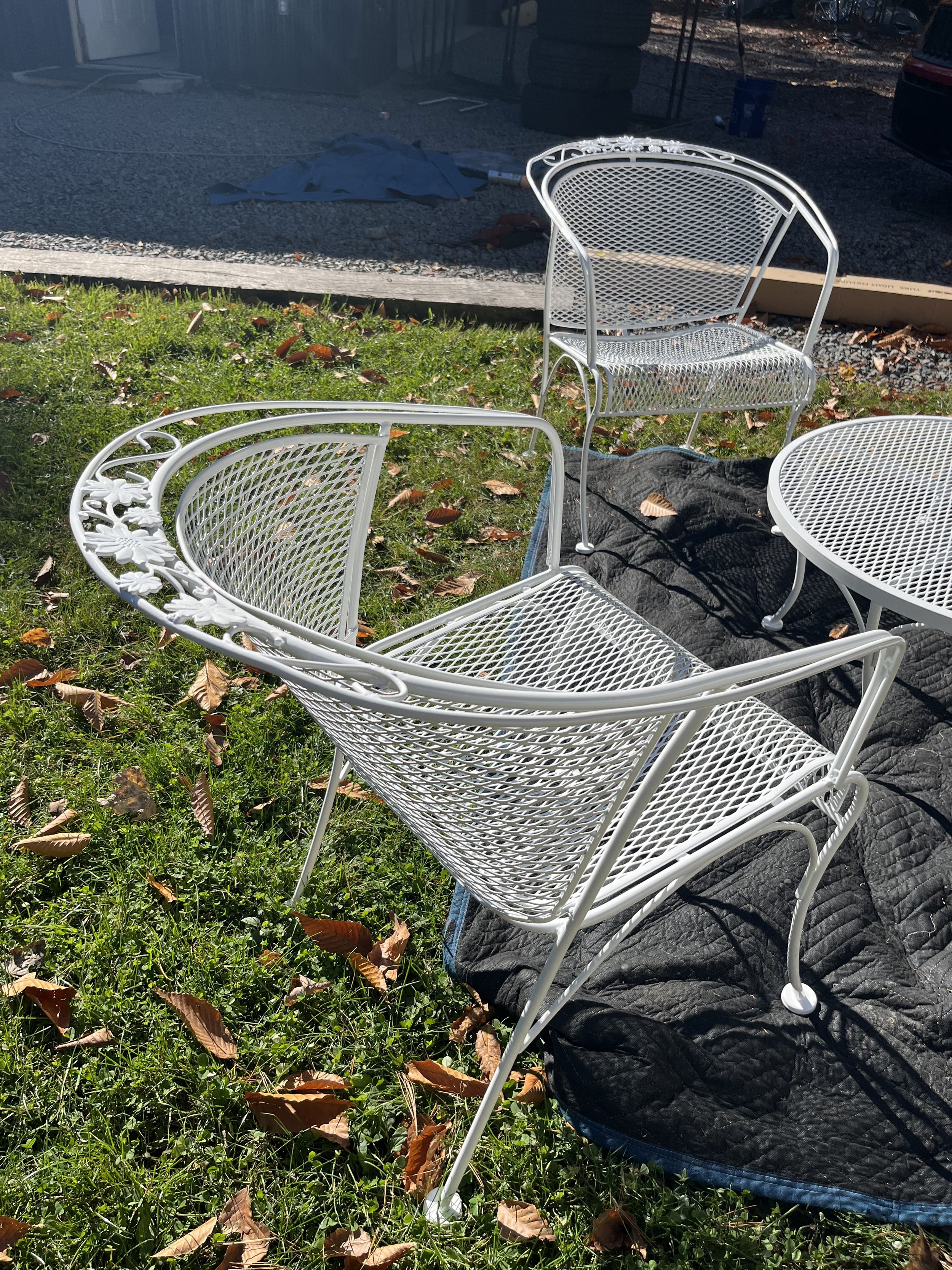 Set of white metal outdoor furniture, including a table with a lattice top, a high-backed chair, and a lounge chair, outdoors on grass with fallen leaves.
