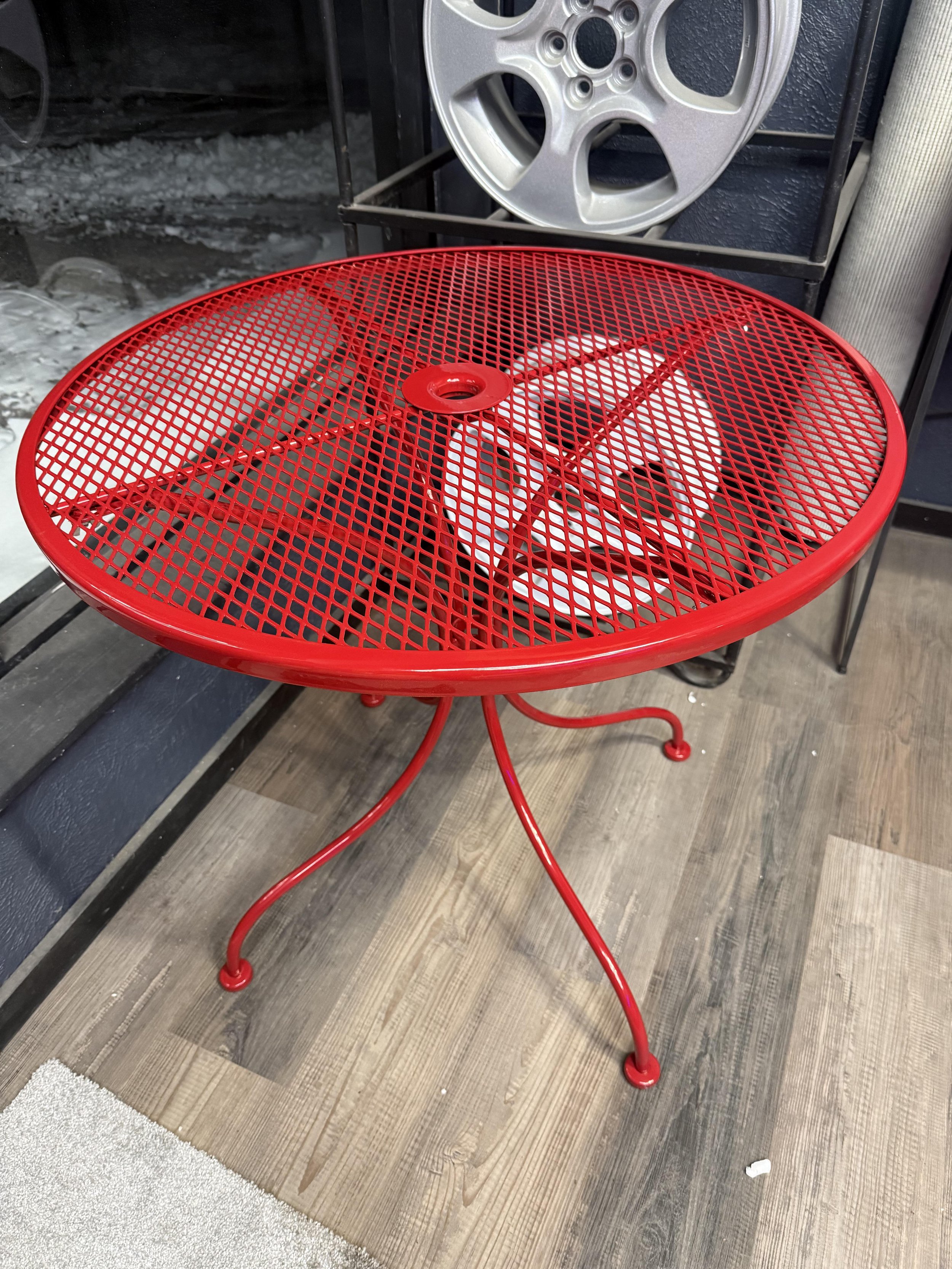 Red metal outdoor table with a mesh top and curved legs, placed on a wooden floor.