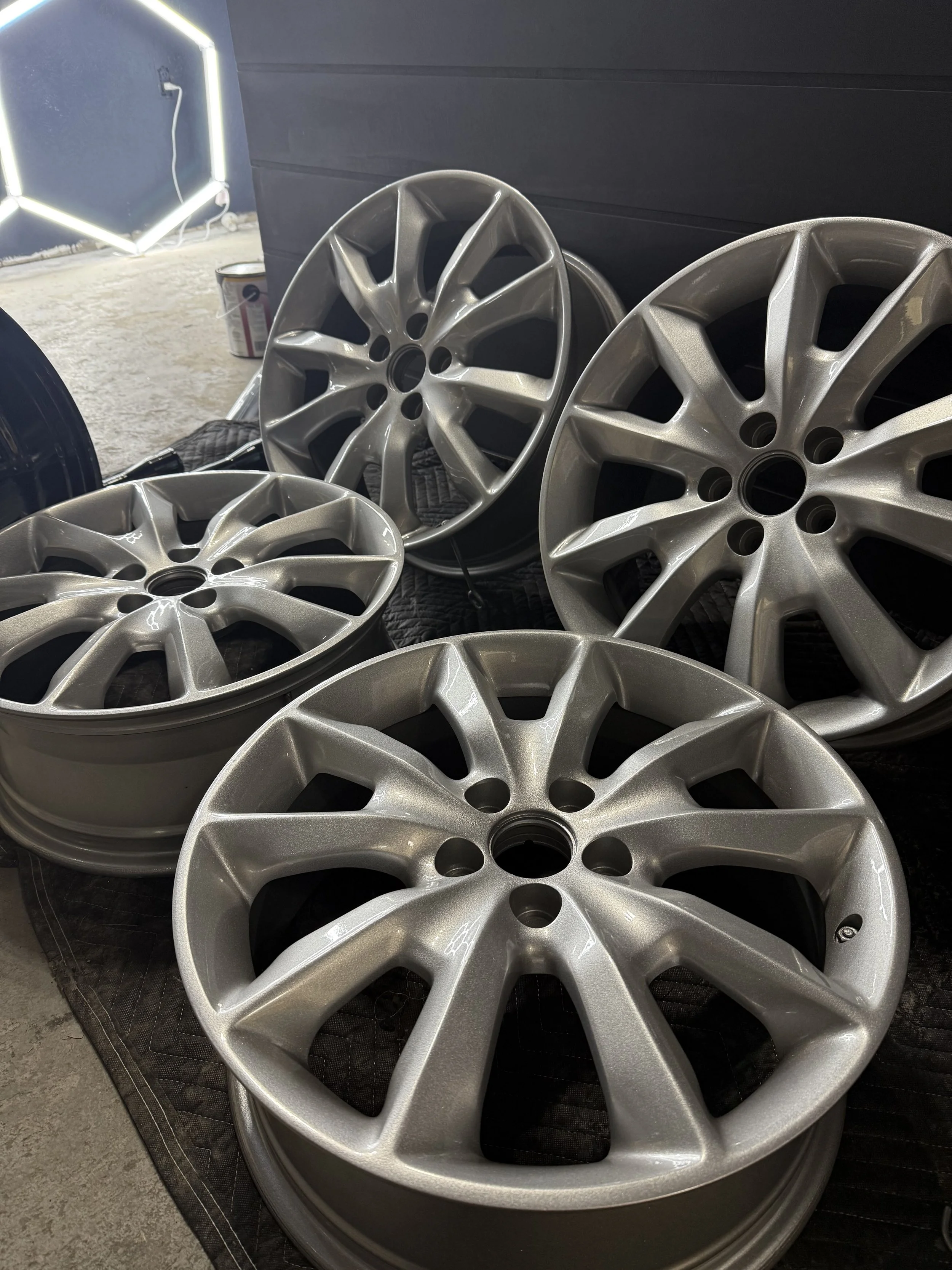 Set of five silver alloy wheels arranged on a black quilted mat, with a dark background and a neon sign partially visible in the background.
