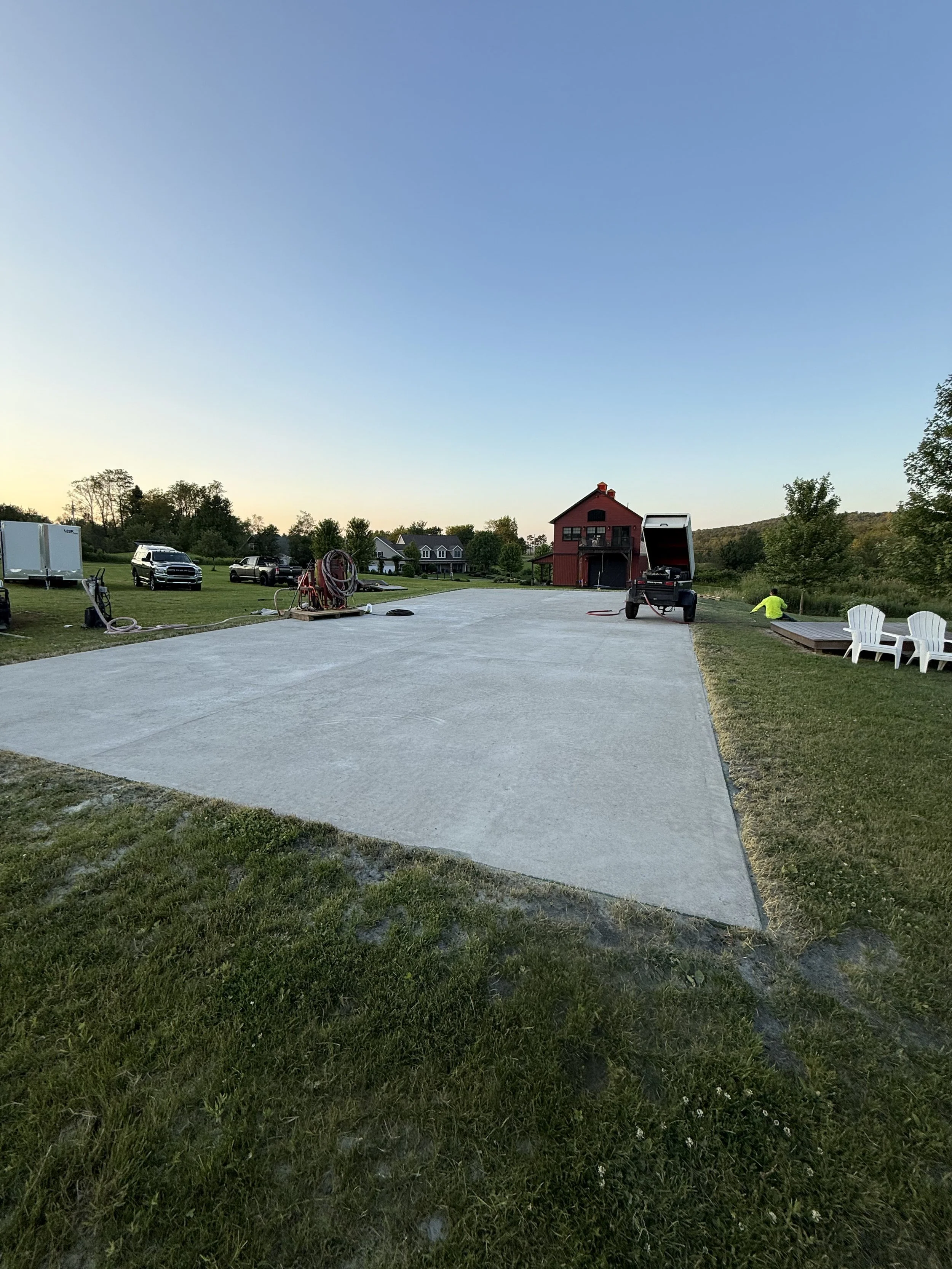 A newly poured concrete slab in a rural backyard with a red barn, lawn chairs, and equipment, against a backdrop of trees and houses under a clear sky.
