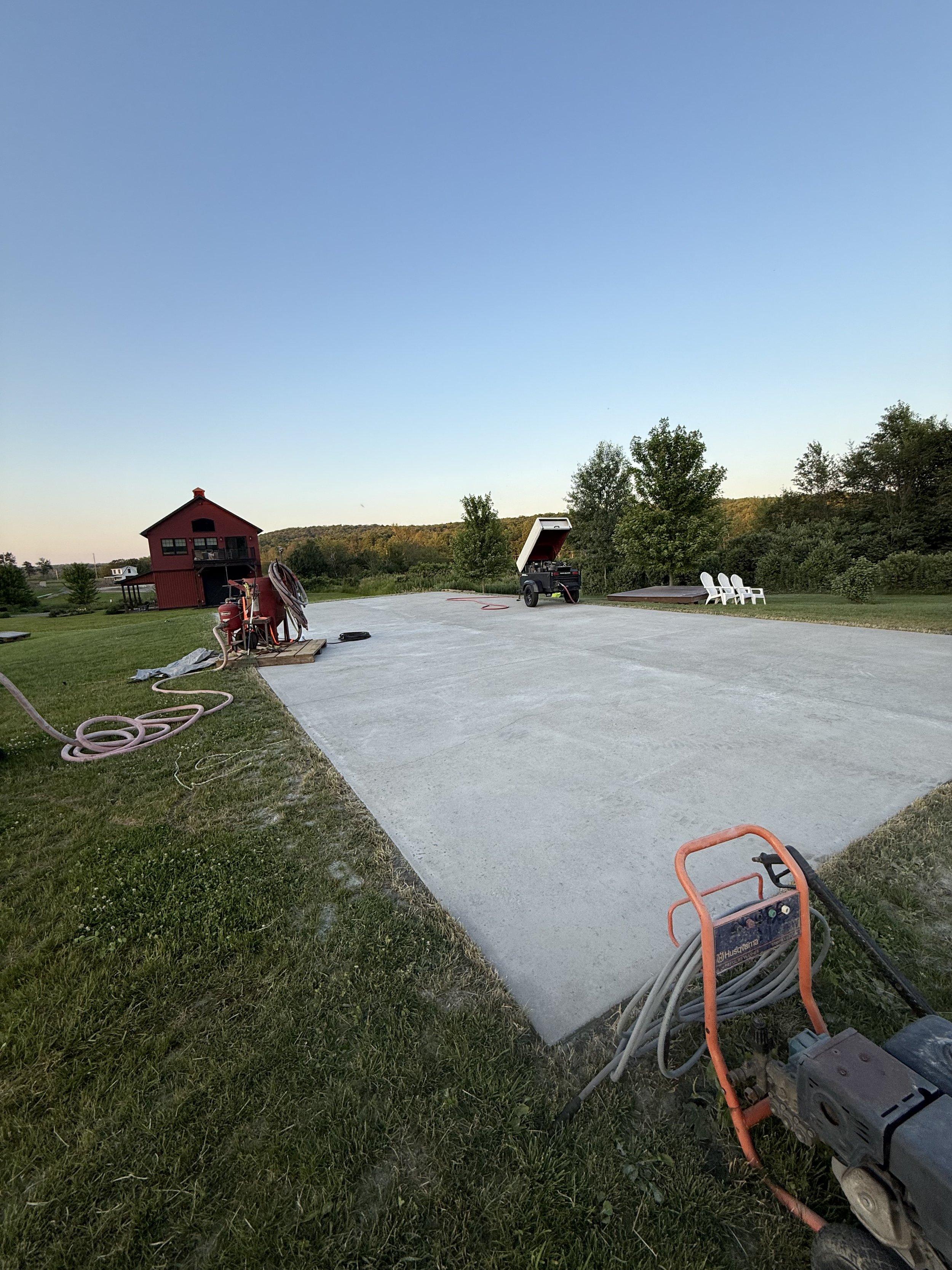 A newly poured concrete patio with construction equipment and hoses, a red barn in the background, and white chairs facing a grassy area surrounded by trees.