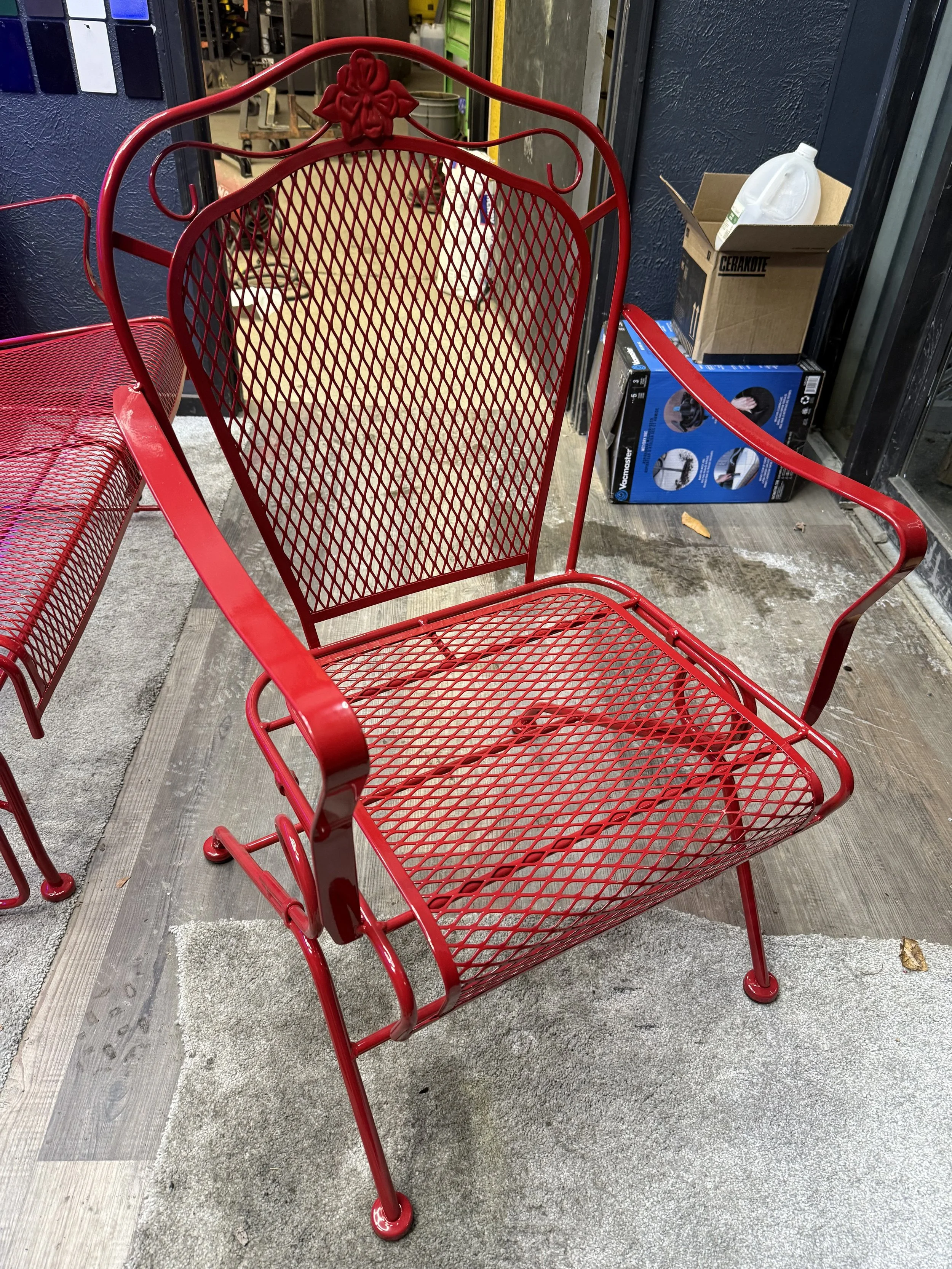 Red metal bench with a decorative backrest featuring a flower in the center, placed outside on a concrete floor.