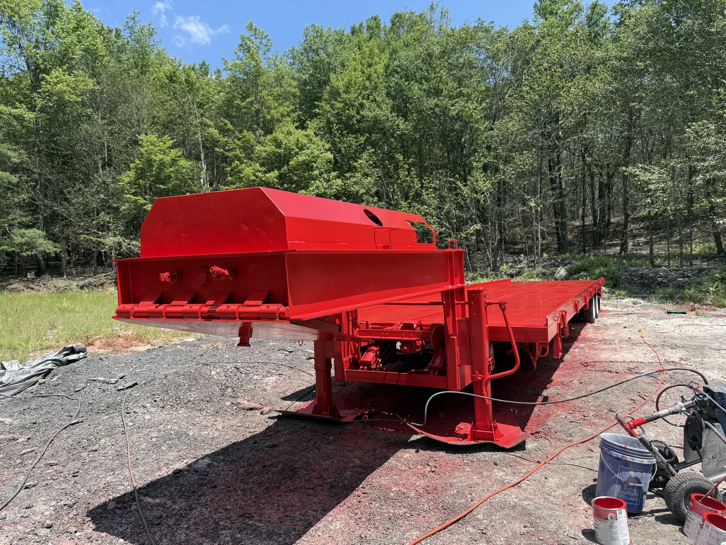 A large red flatbed truck trailer is set up outdoors on a gravel surface, with a forested area and blue sky visible in the background.