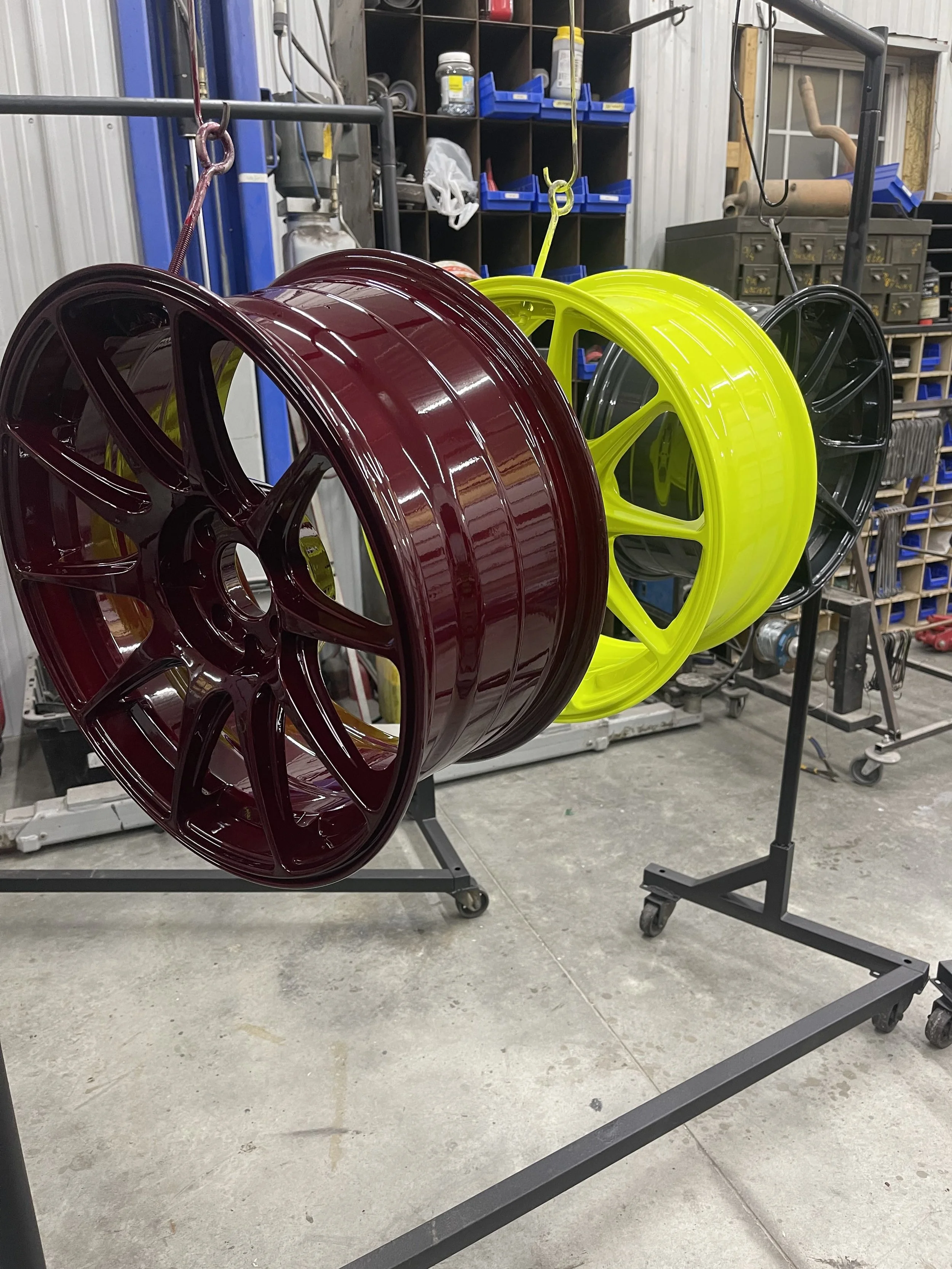Three painted car wheels hanging on a rack inside a workshop, with shelves and tools in the background.