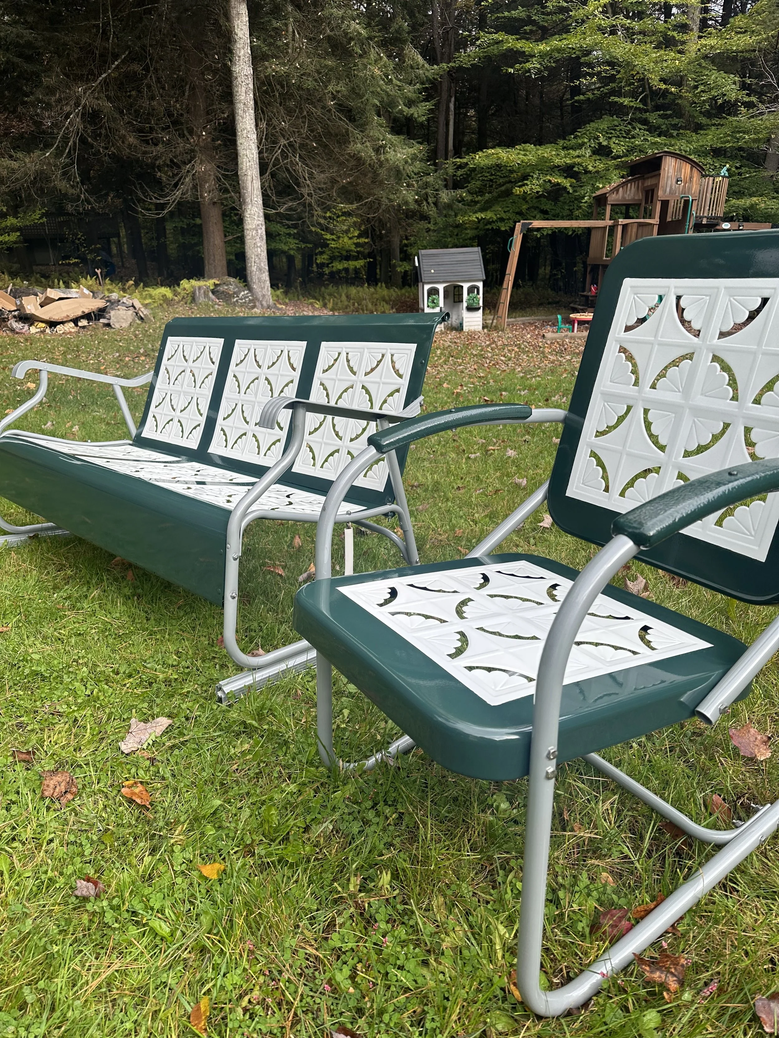 Two outdoor metal and plastic chairs with white decorative backs and seats are placed on green grass in a backyard, with a playhouse and trees in the background.