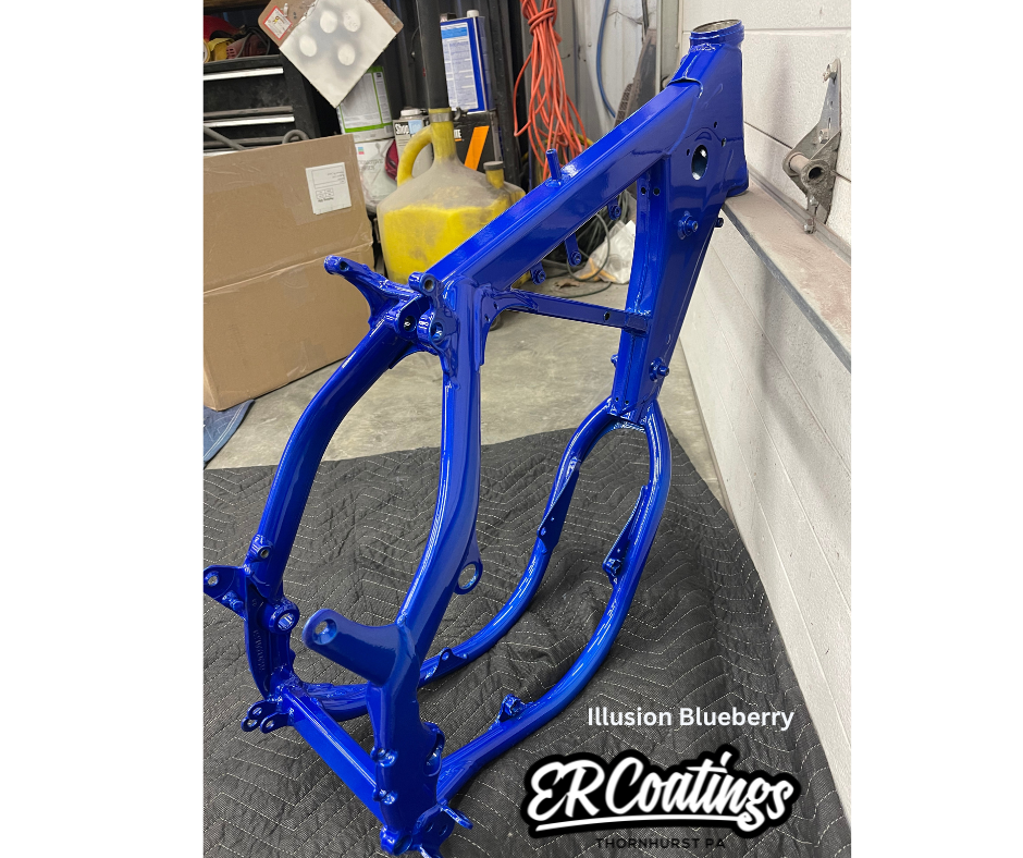 A blue painted motorcycle frame in a garage or workshop setting with various containers, tools, and cords in the background.