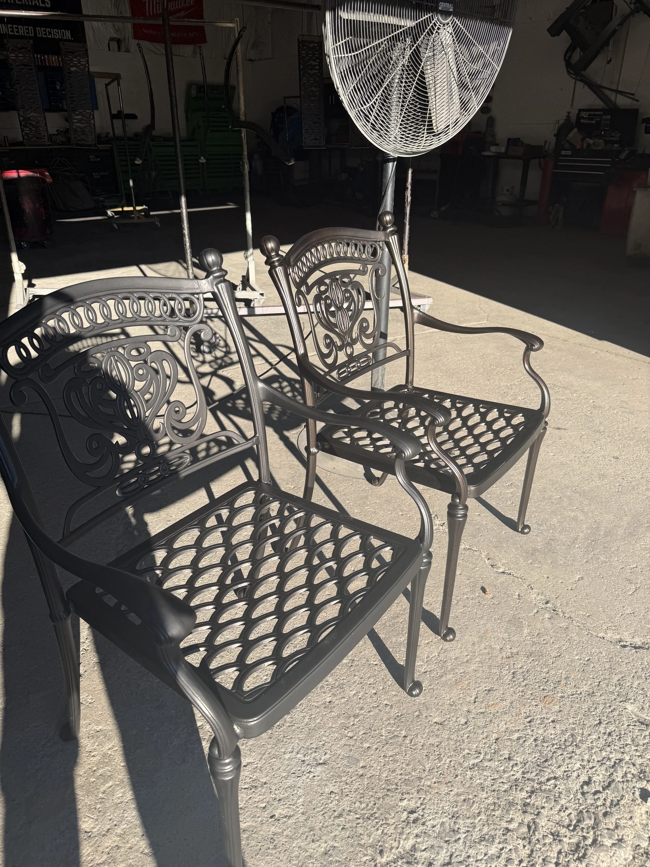 Two black metal outdoor chairs with decorative backs and seats, placed on a concrete surface outdoors, with a large fan and workshop in the background.
