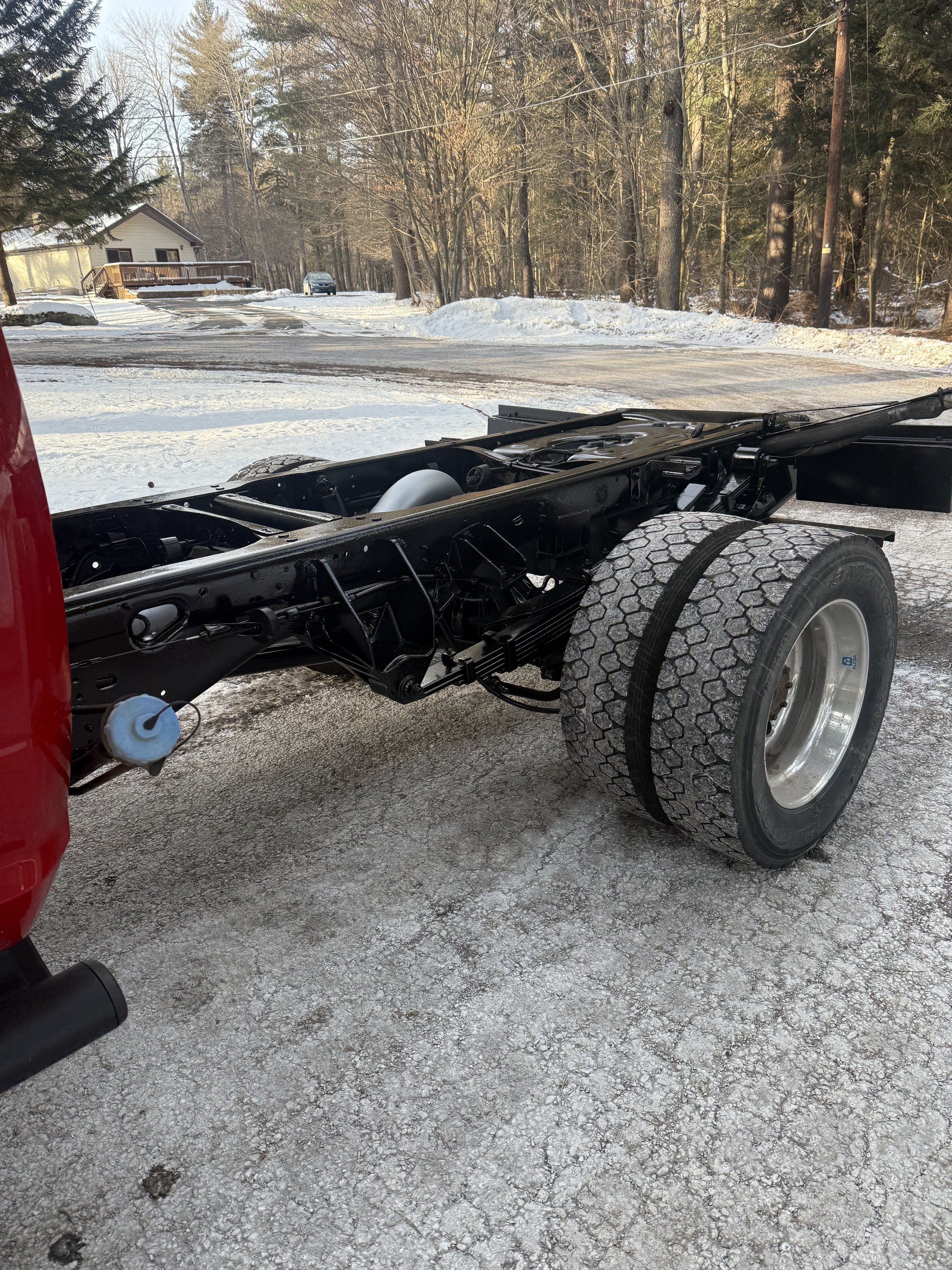 Image shows the chassis of a truck on a snowy ground with a road, houses, trees, and a car in the background.