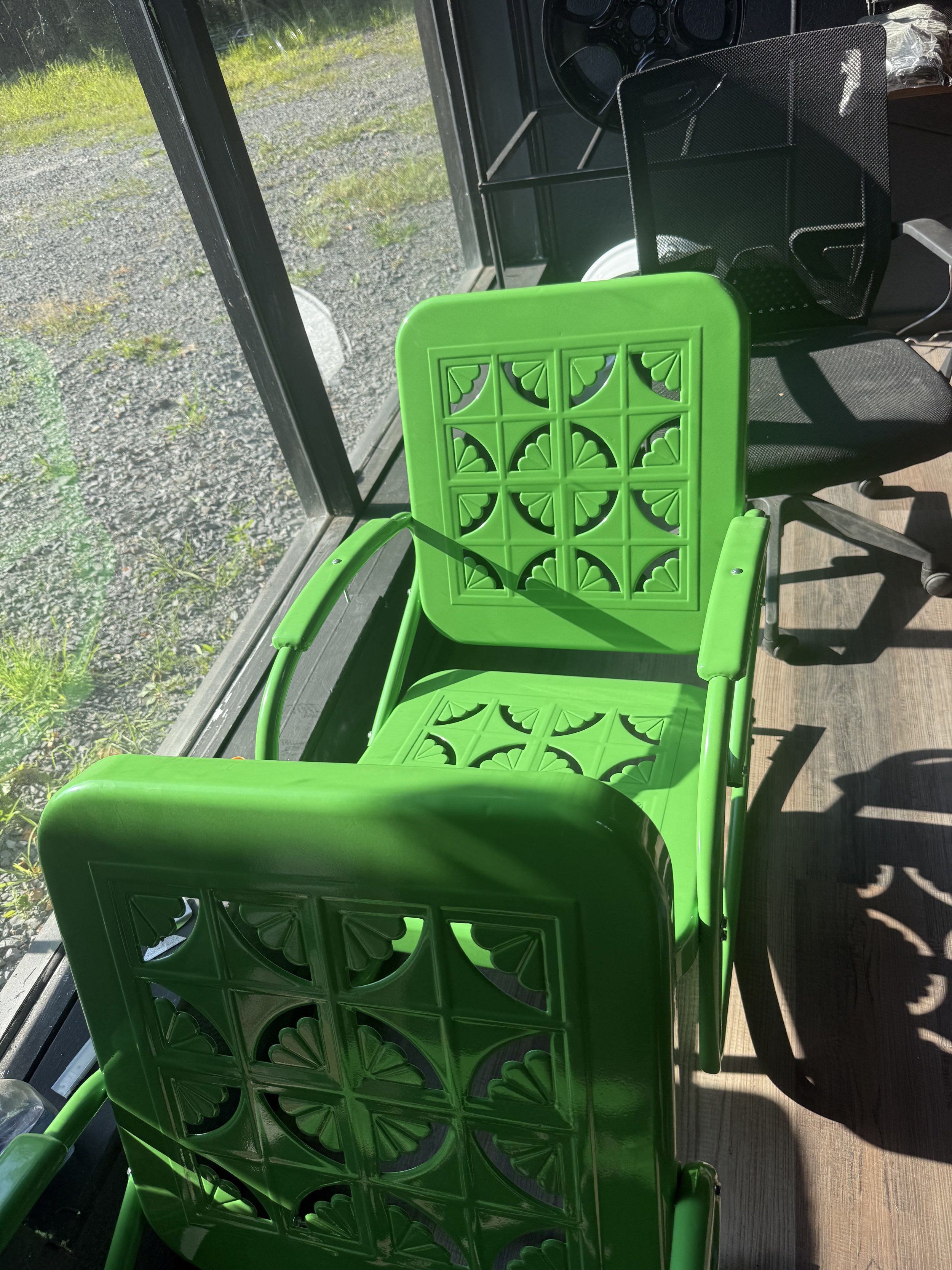 Green lawn chairs with decorative cutouts, placed inside near a glass window, casting shadows on wooden floor.