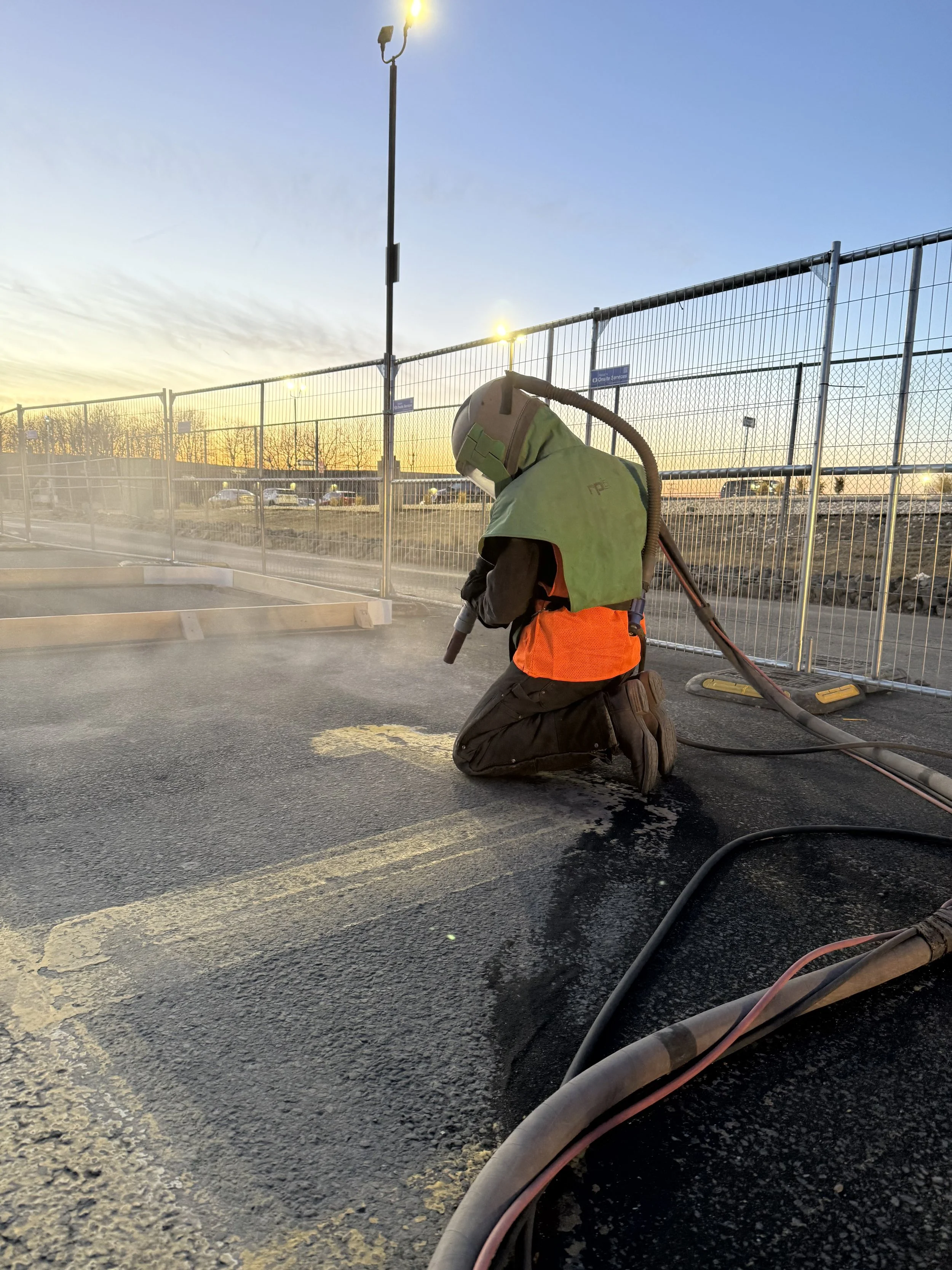A worker wearing a helmet, safety vest, and kneeling on the ground, using equipment to asphalt or patch a road during sunset, with a fence and parking lot in background.