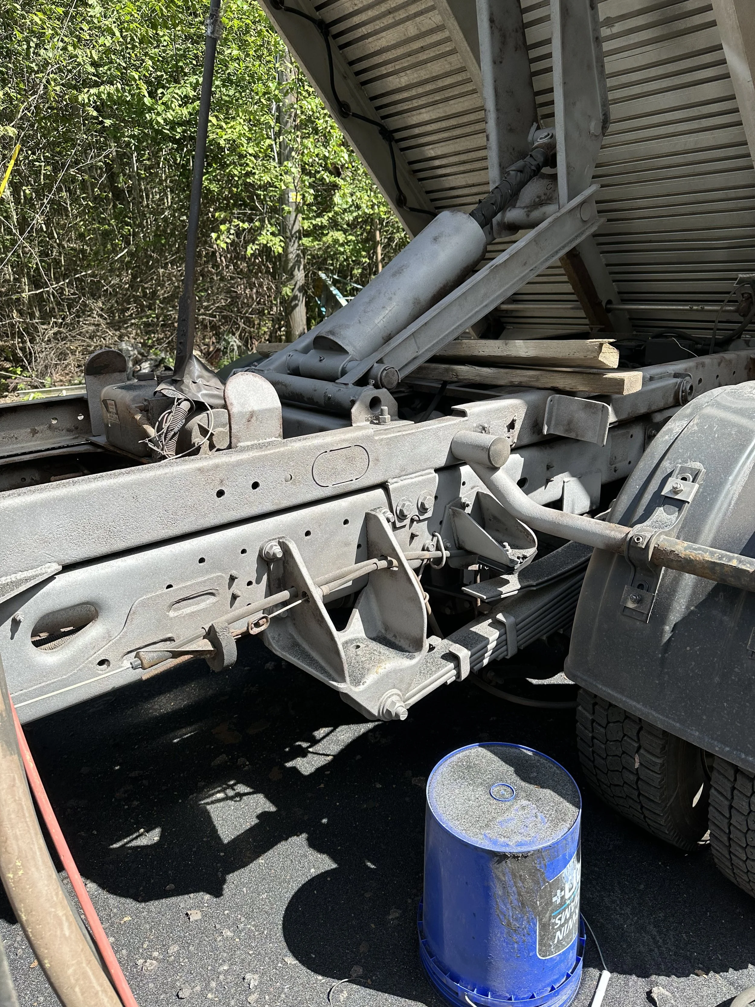 Close-up of a mechanical structure, likely part of a vehicle, with hydraulic cylinders and metal framing, on a paved surface with a blue bucket nearby, and green foliage in the background.