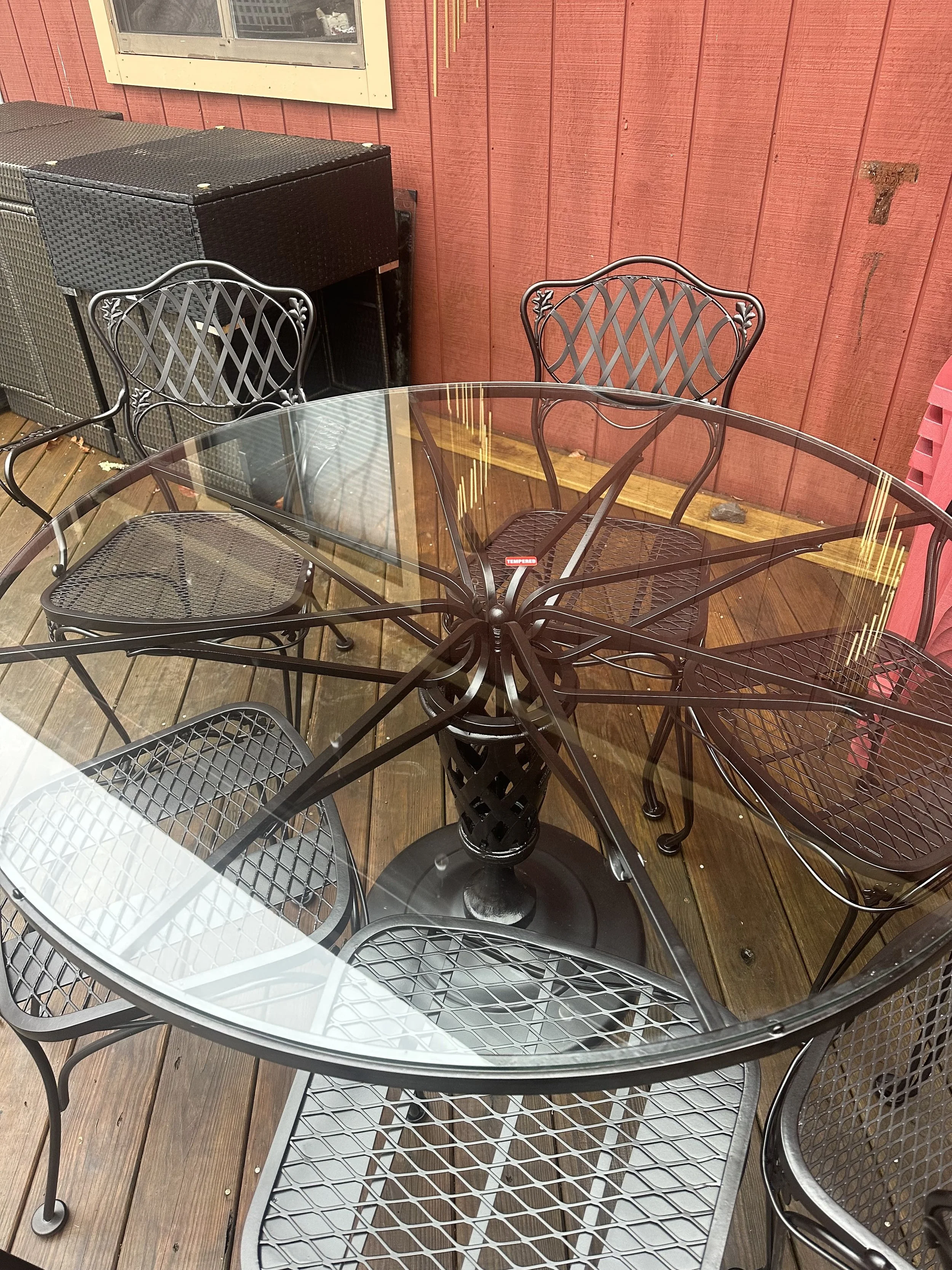 A round glass outdoor table with five black metal chairs around it on a wooden deck, with a red wooden wall in the background and a window.