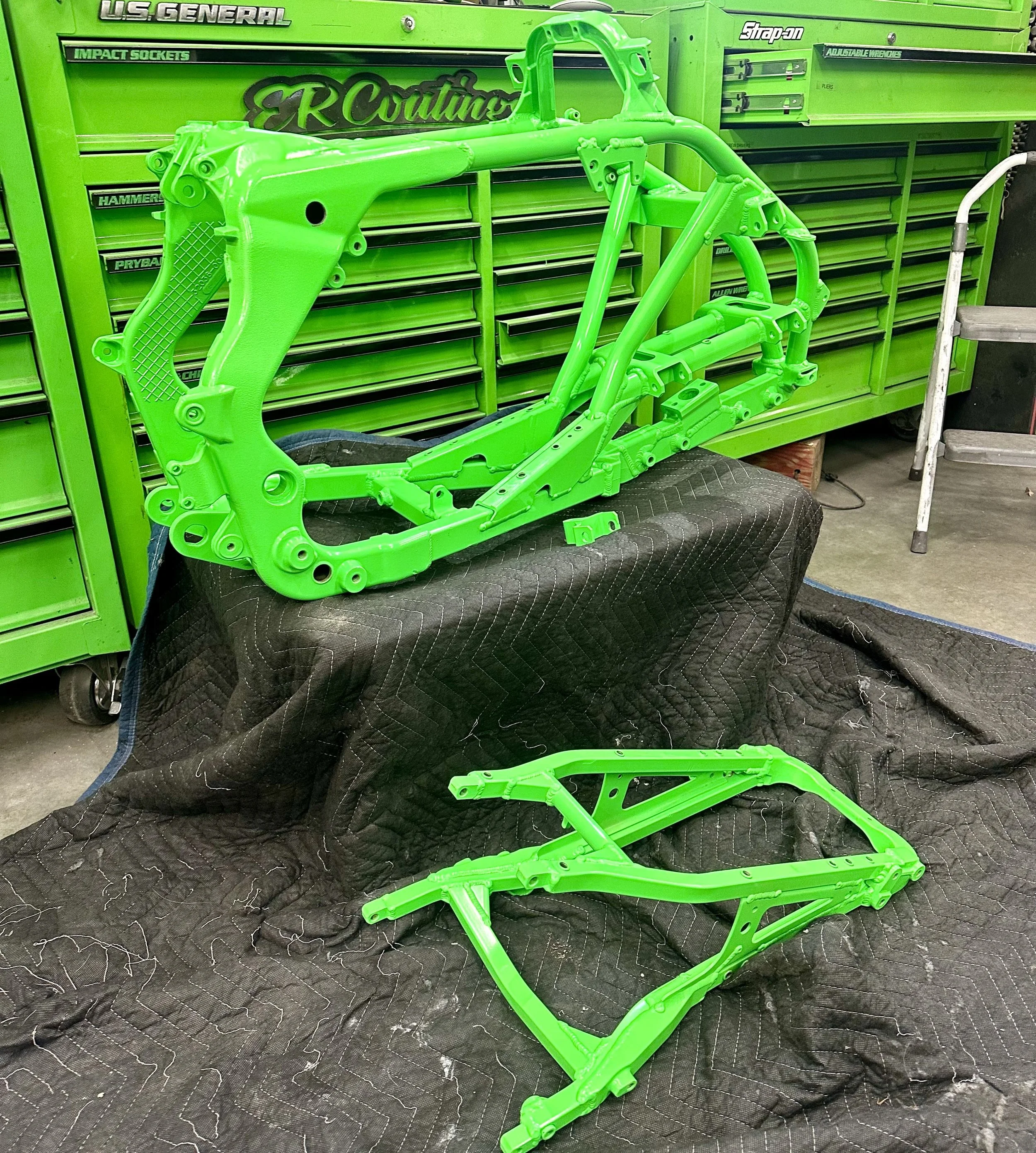 Bright green motorcycle frame parts on a black cloth in a workshop with green tool cabinets in the background.