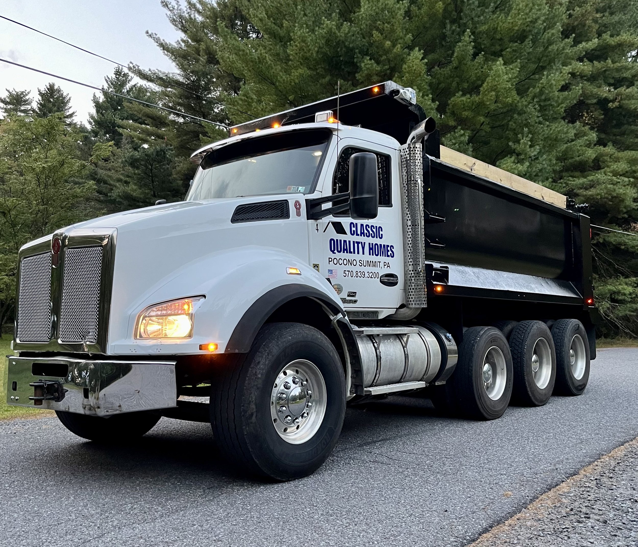A large white and black dump truck parked on a road with trees in the background. The truck has the text 'Classic Quality Homes' and 'Pocono Summit, PA' on the side.