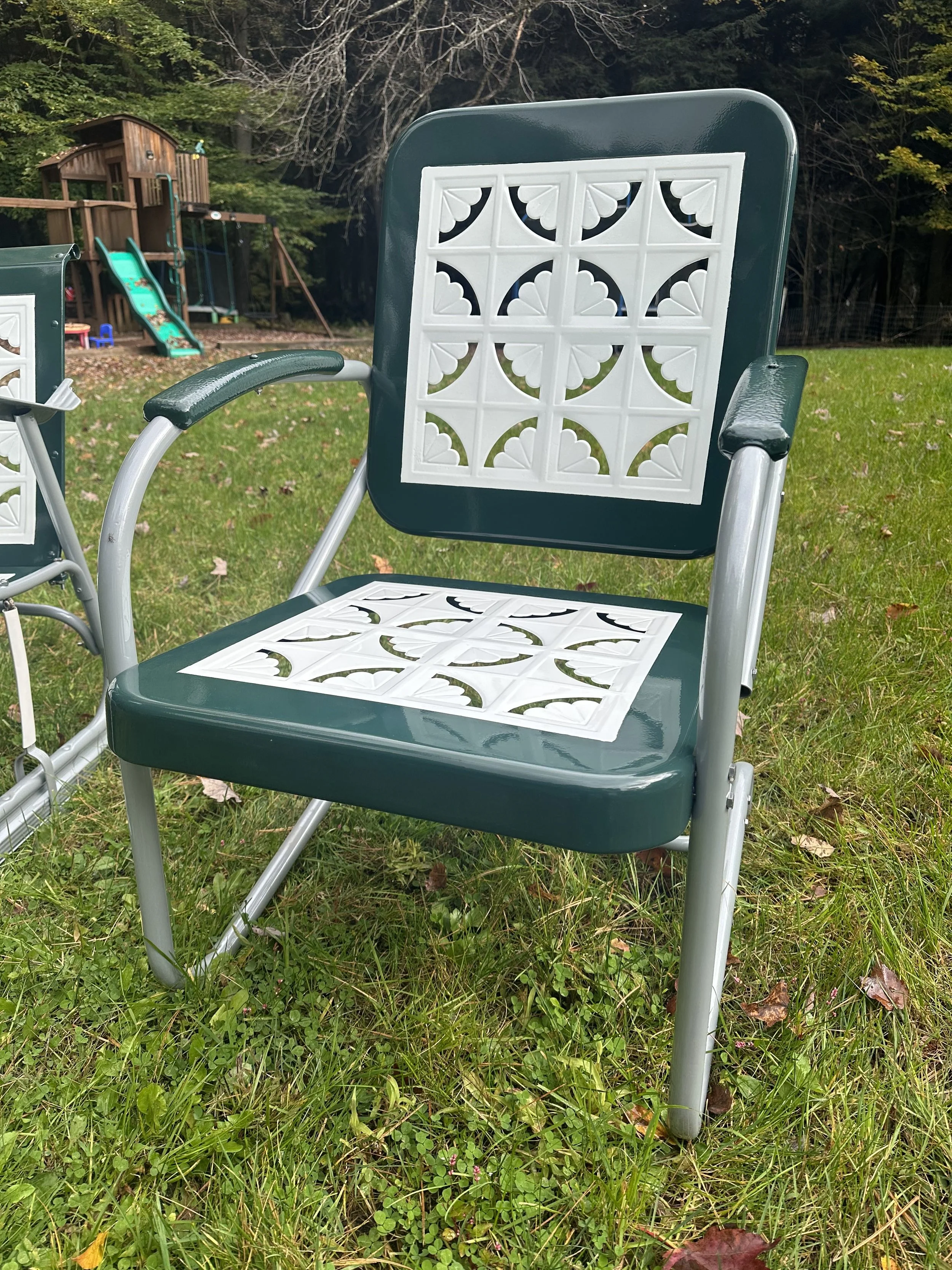 A green and white vintage-style outdoor chair with decorative white panels on the backrest and seat, on a grassy lawn near a playground and trees.