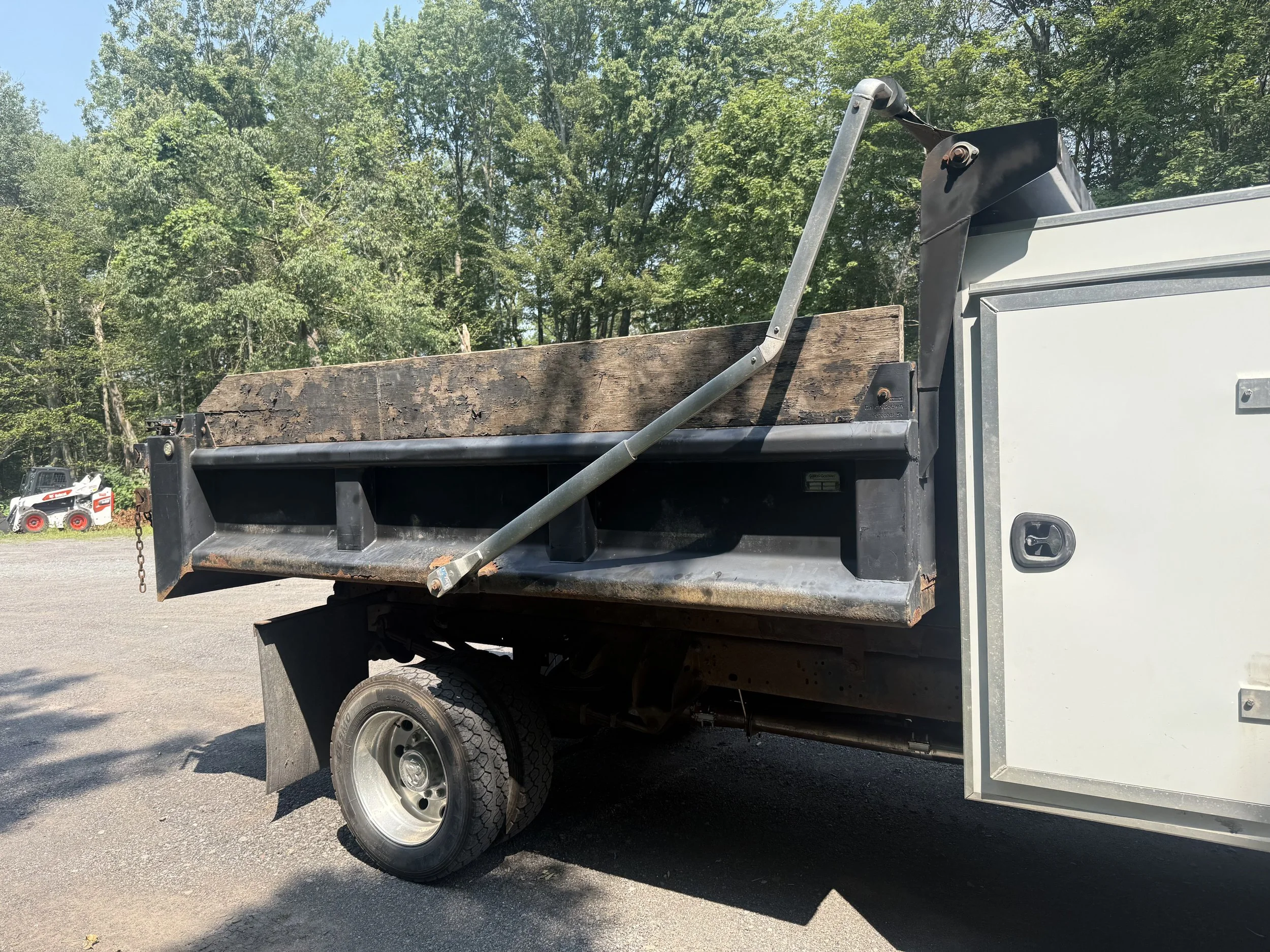 Side view of a utility truck bed with a wooden plank, hydraulic arm, and black container, parked on a gravel surface with green trees in the background.