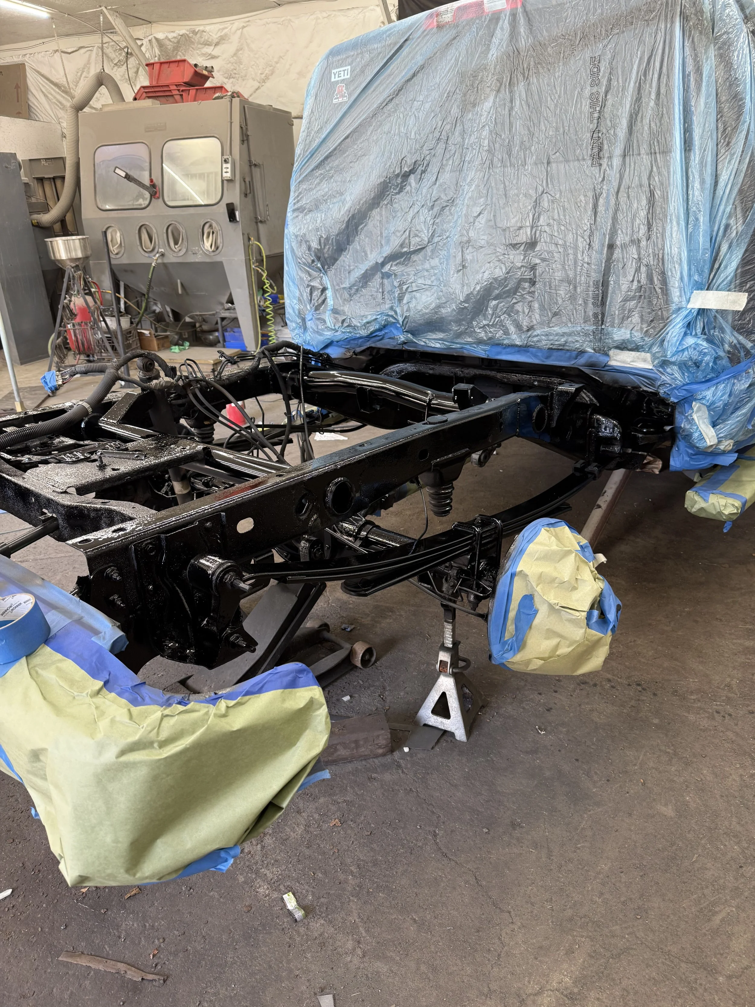 Vehicle being painted or assembled in a workshop, with parts covered in masking paper and tape, and a blue tarp covering some sections.