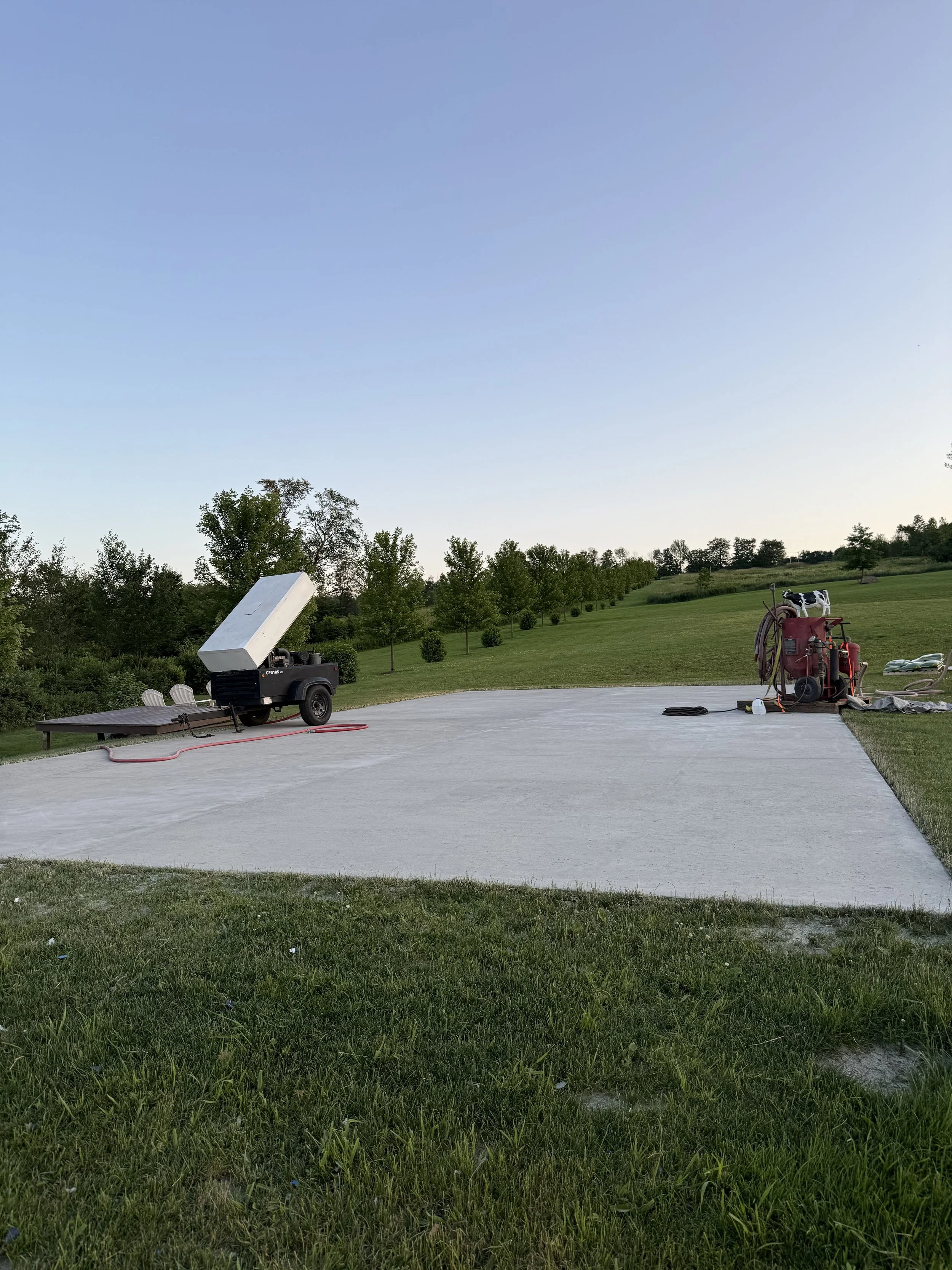 A concrete slab with construction equipment, including a portable generator and other tools, in an outdoor grassy area with trees and open sky in the background.