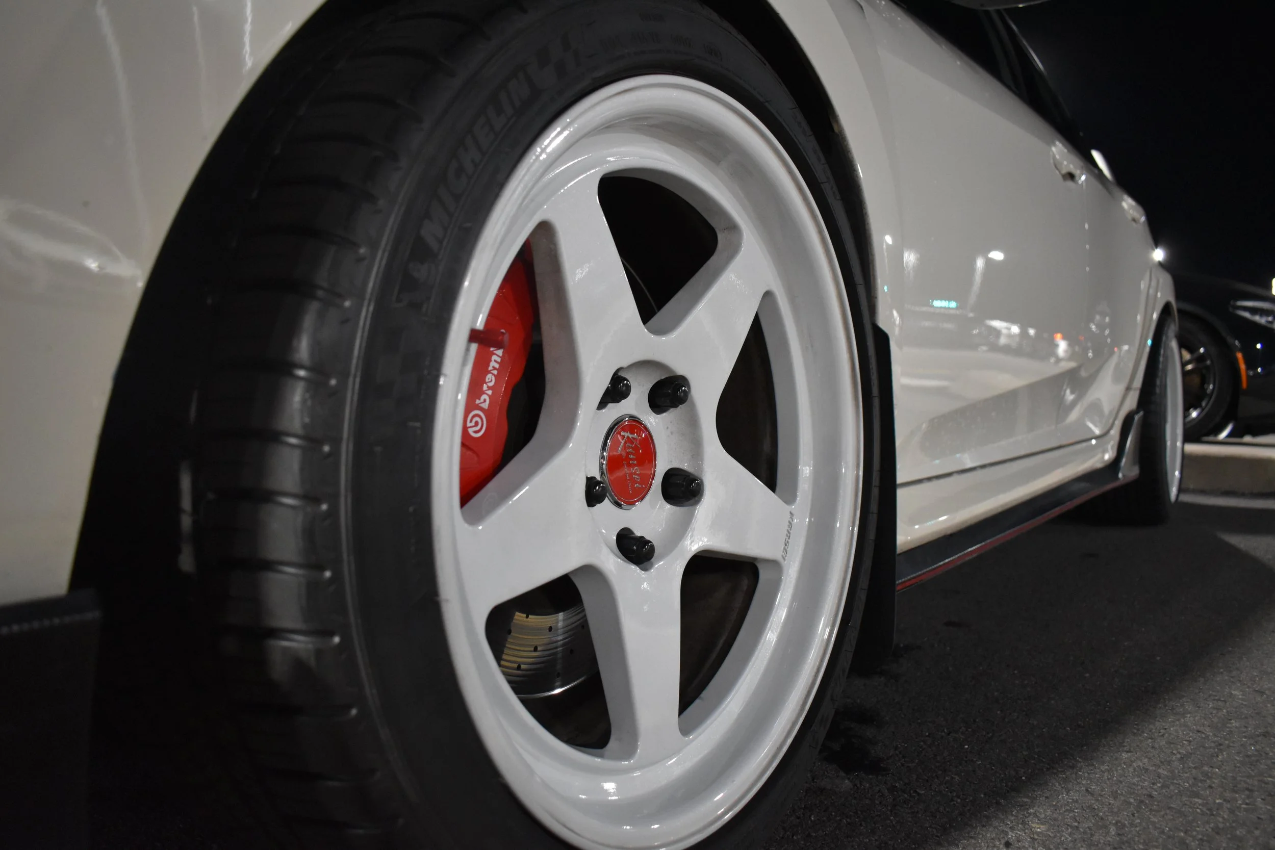 Close-up of a white car's front wheel showing a white rim with black bolts, red brake caliper, black tire, and part of the vehicle body parked at night.