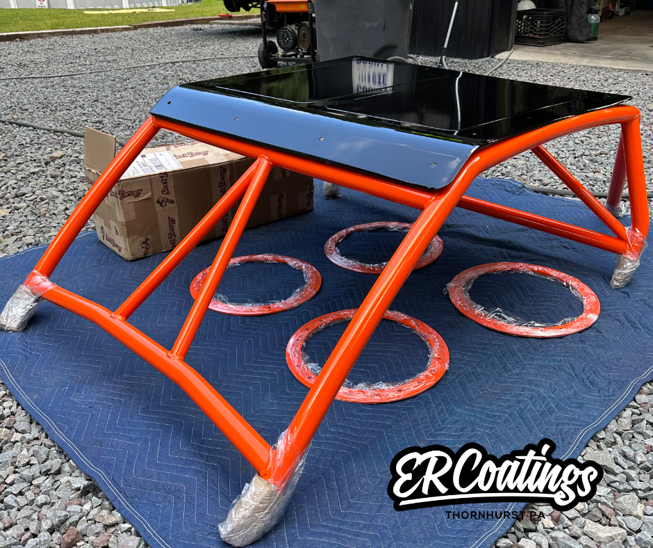 A racing car roll cage frame with a black flat panel attached to the top, orange painted metal tubing, and four red circular rings on the ground inside a garage. The frame's ends are covered with plastic protectors, and a cardboard box is nearby. The