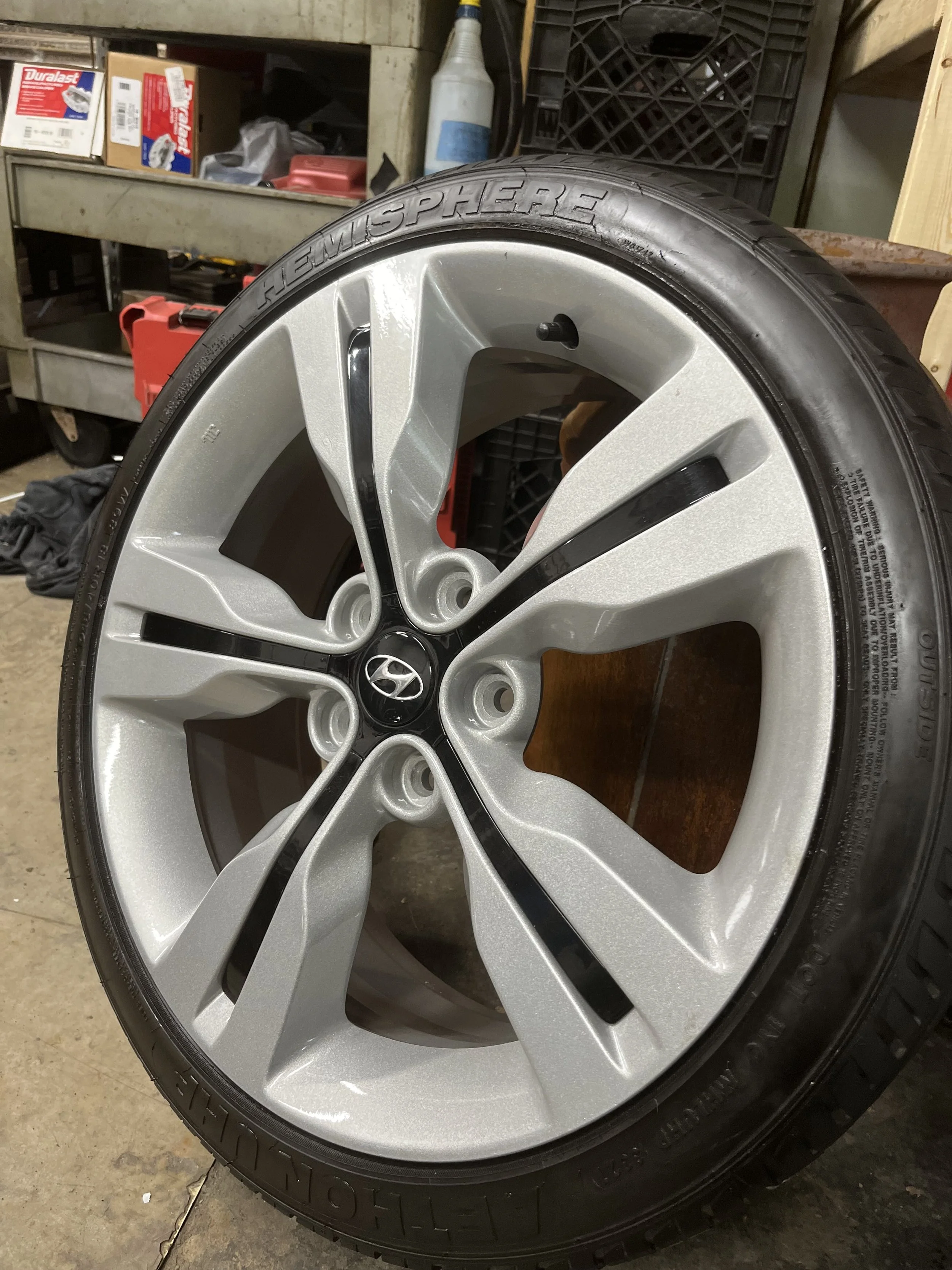 A silver five-spoke Hyundai alloy wheel with a black Hyundai center cap, mounted on a Bridgestone Ecopia tire, placed on a concrete floor in a garage or workshop environment.