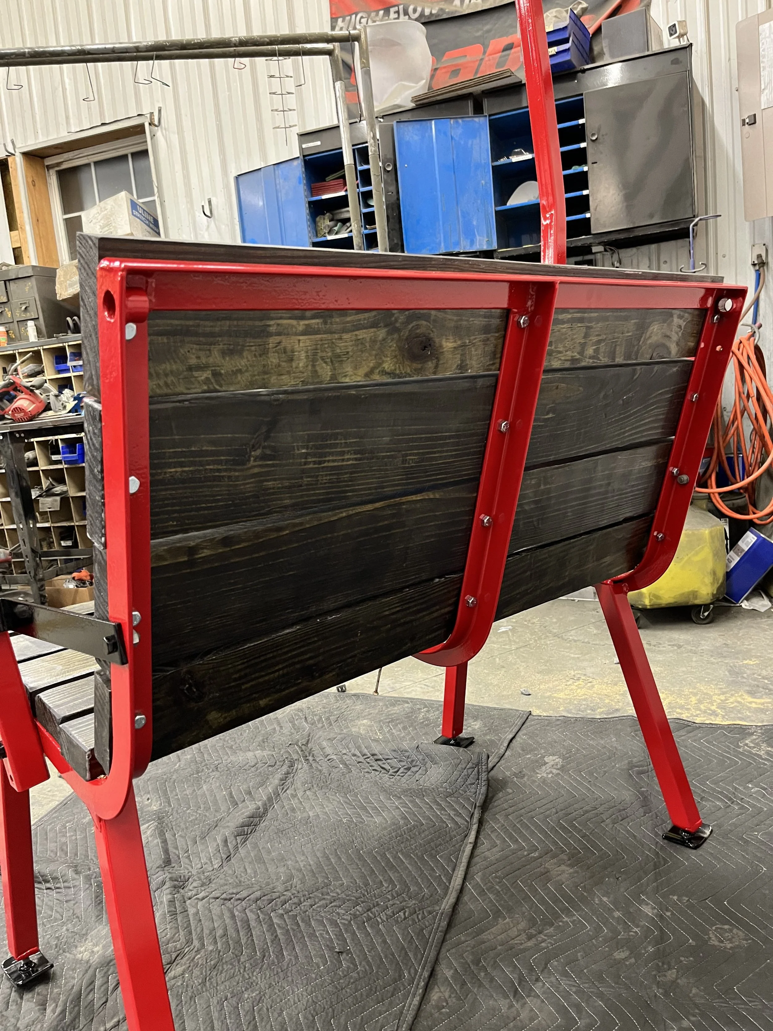 A wood-fired pizza oven frame with red metal edges and black exterior panels, on red metal legs, in a workshop setting with tools and storage cabinets in the background.
