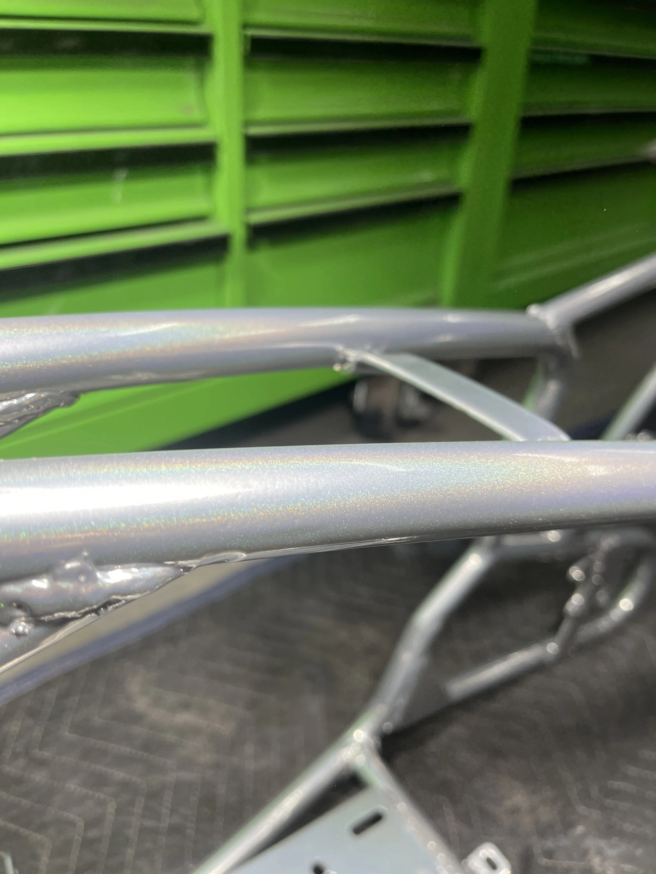 Close-up of metal rails with a shiny, iridescent finish, part of a bicycle or cart, with green shelves in the background.