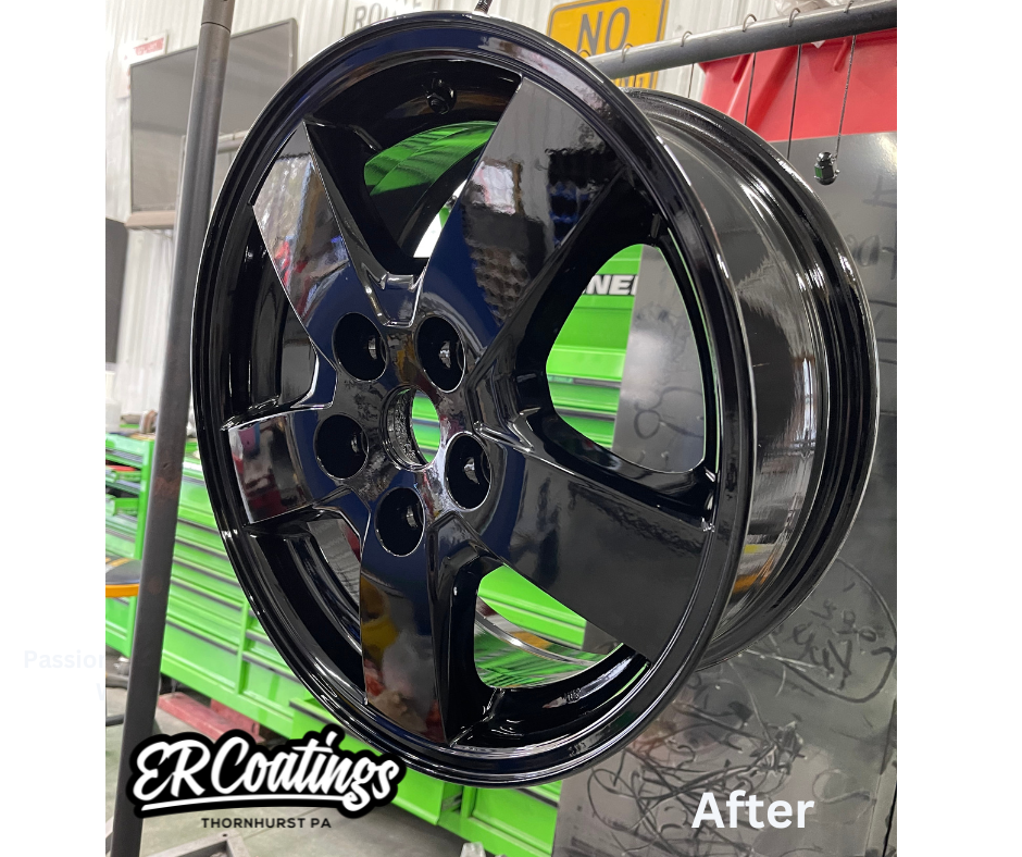 A black, shiny car wheel rim with a modern five-spoke design, mounted on a display stand at a workshop, with a background of green tool drawers and workshop equipment.
