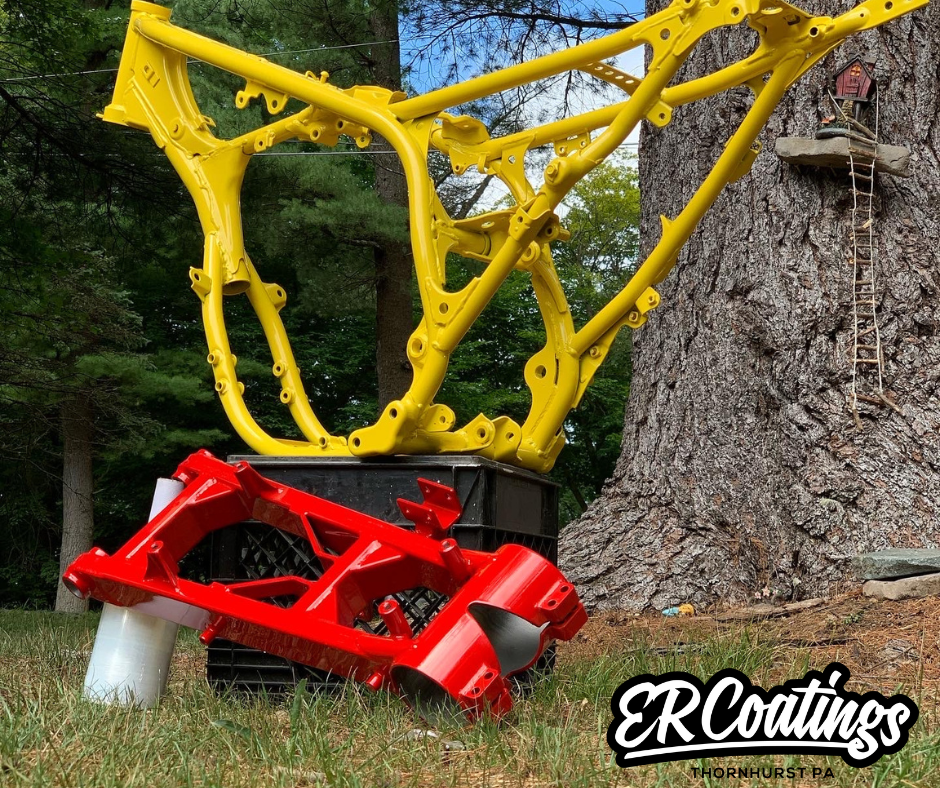 Disassembled yellow motorcycle frame, red suspension components, and black crate near a large tree with a birdhouse, outdoors with green trees in the background.