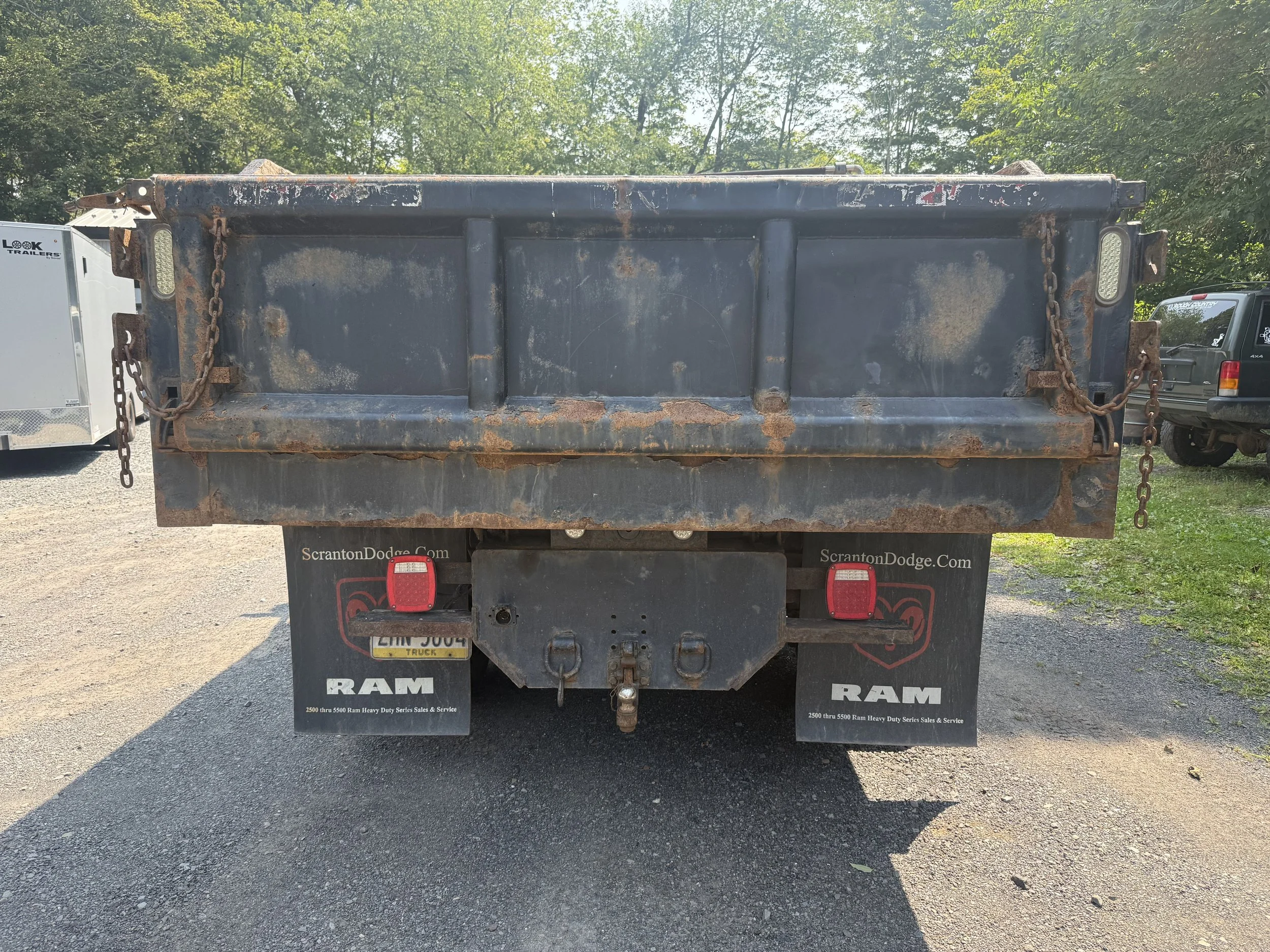 Back view of a rusty black flatbed truck with chains on the sides, parked on gravel. Behind, trees and some other vehicles are visible.