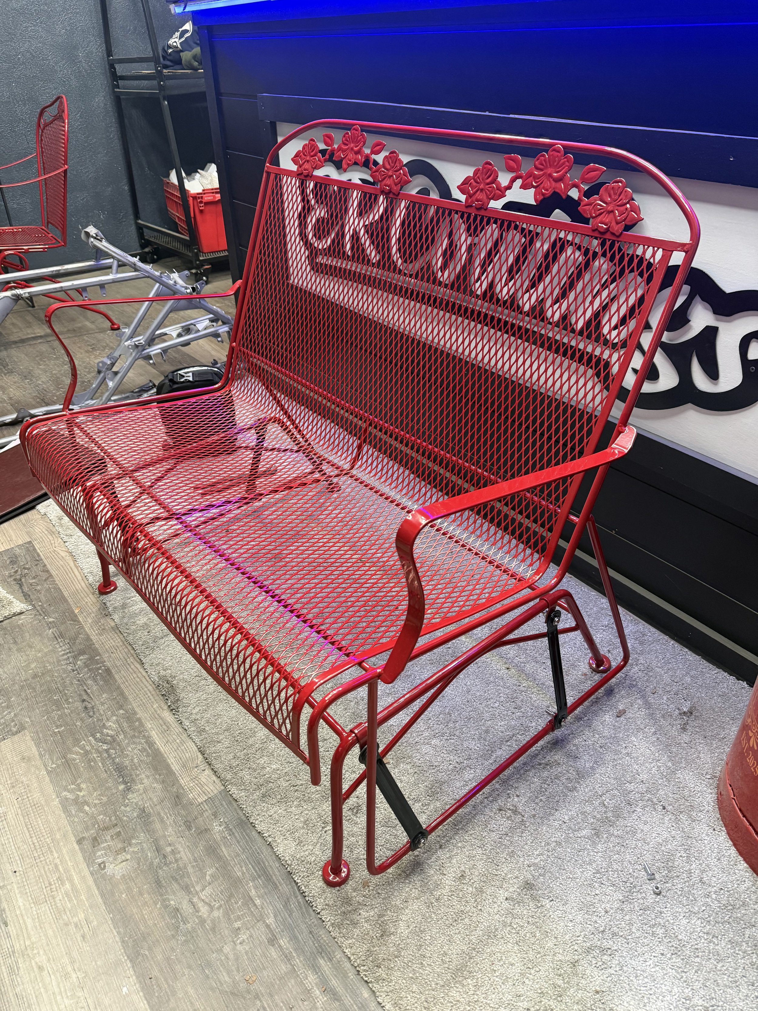 Red metal outdoor bench with floral details on the backrest, placed on a beige rug inside a store with a dark wall and other furniture in the background.