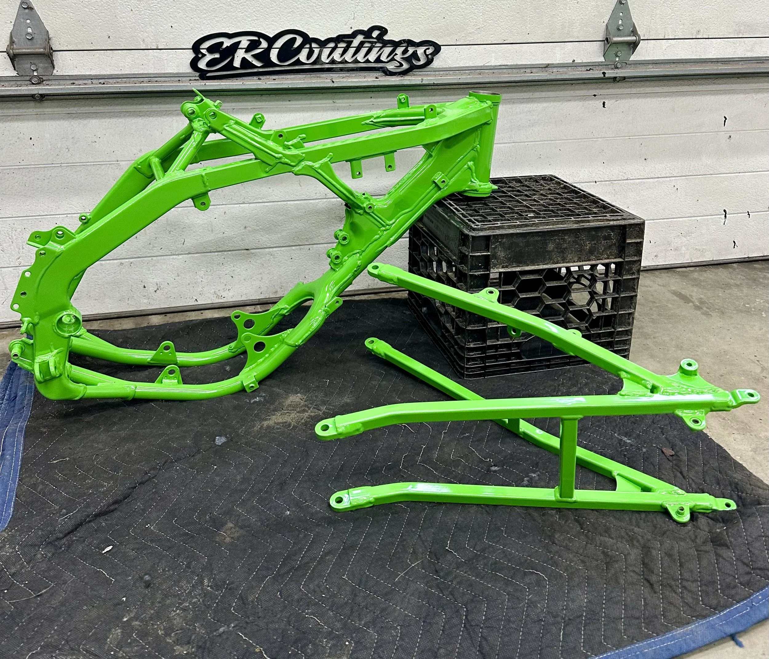 Bright green motorcycle frame parts on a black tarp in a workshop with a white garage door and a black sign that reads 'ER Customs' in the background.