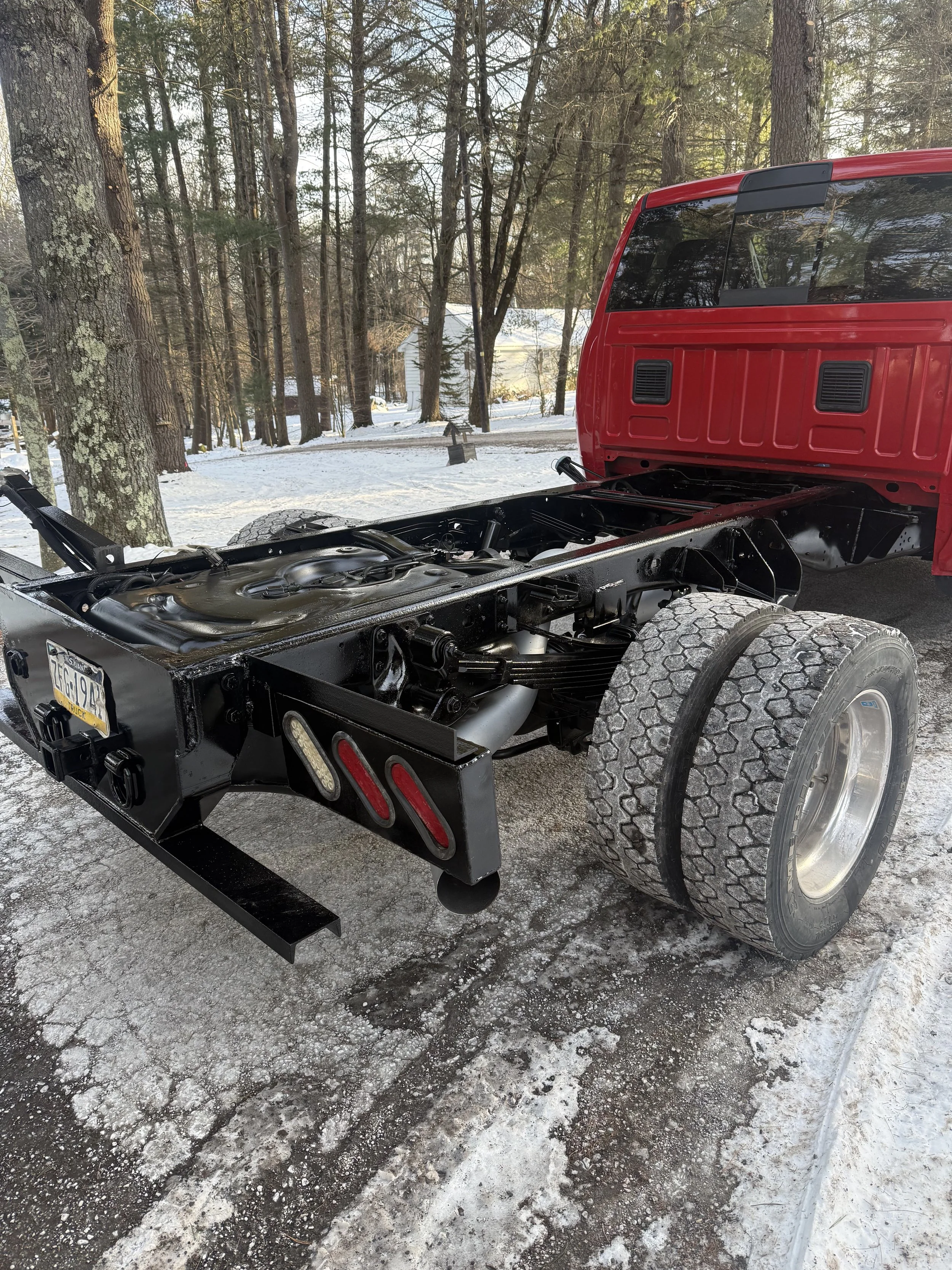 Red truck with no bed, showing the chassis and rear wheels, parked on a snowy ground with trees in the background.
