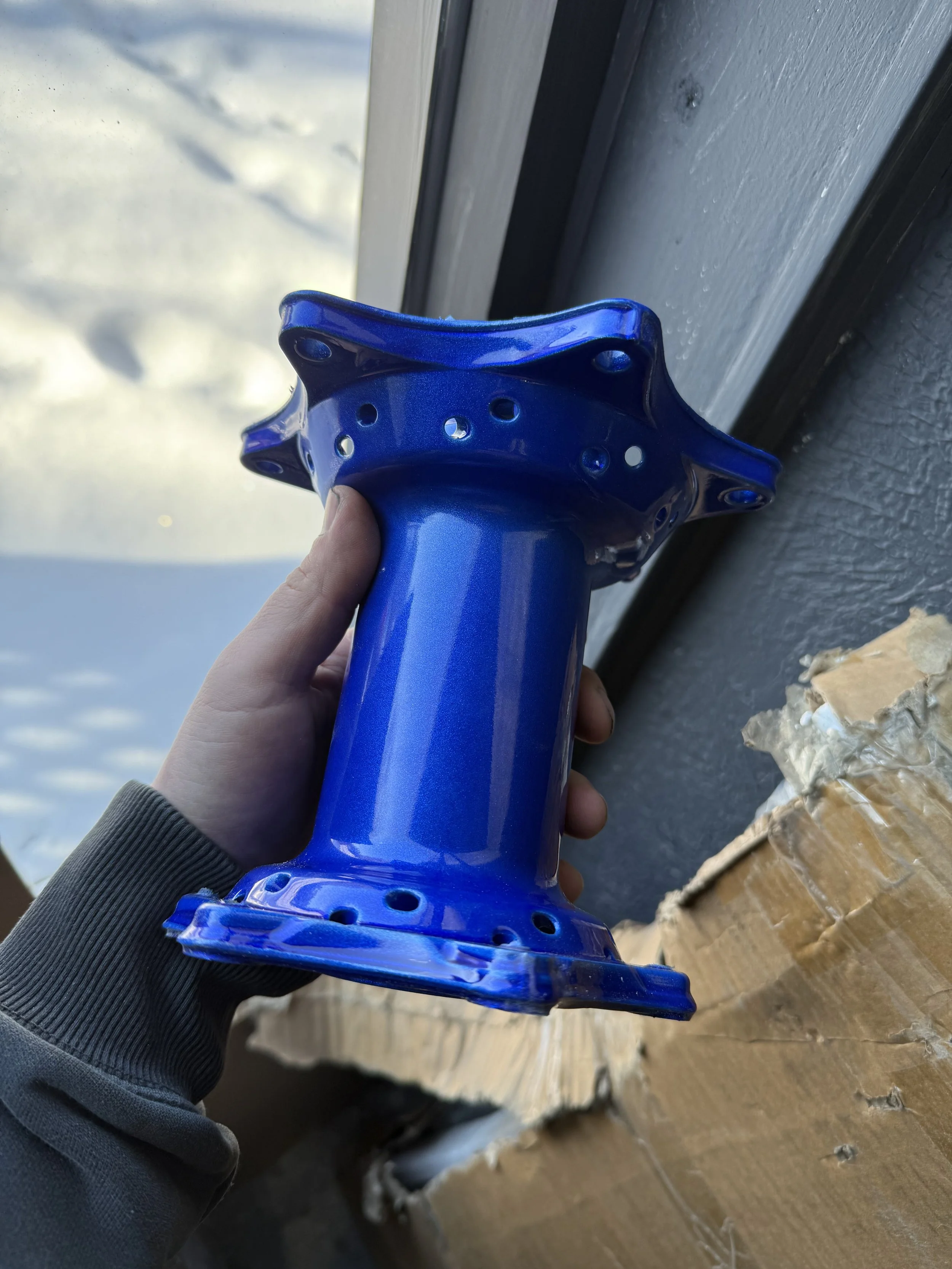 A person holding a blue ceramic object with multiple holes, likely a part of a pottery piece or a decorative item, near a window with snow visible outside.