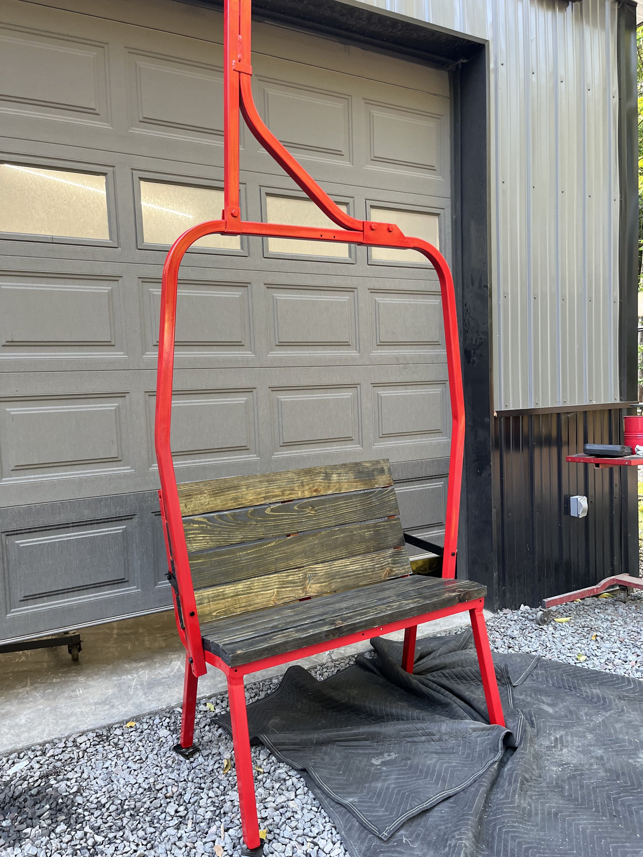 A red outdoor swing with a wooden slat seat and backrest, positioned in front of a gray garage door and black metal siding, on a gravel surface with a black mat underneath.