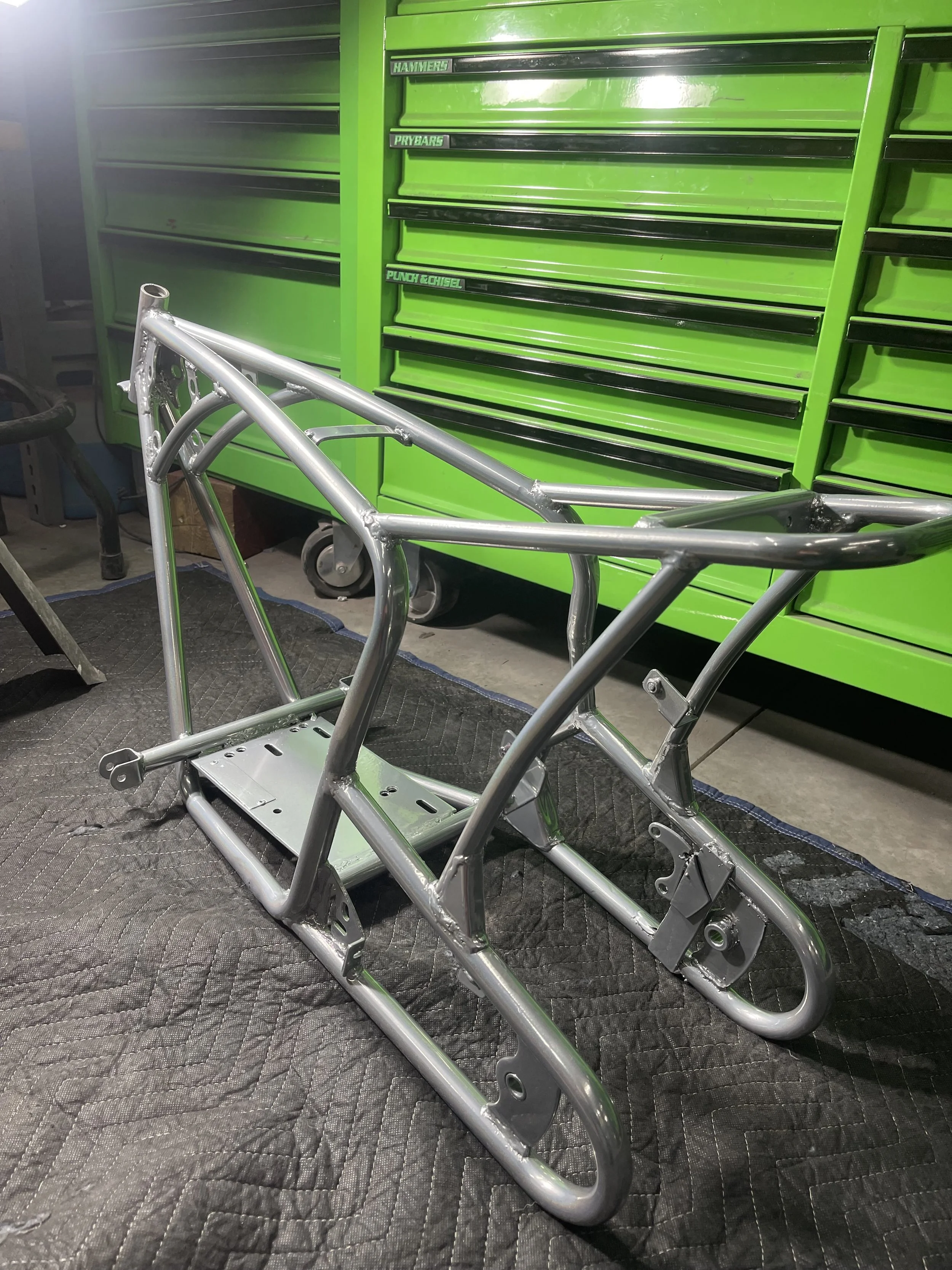 Metal motorcycle frame on a black padded mat in front of a green tool chest in a workshop.