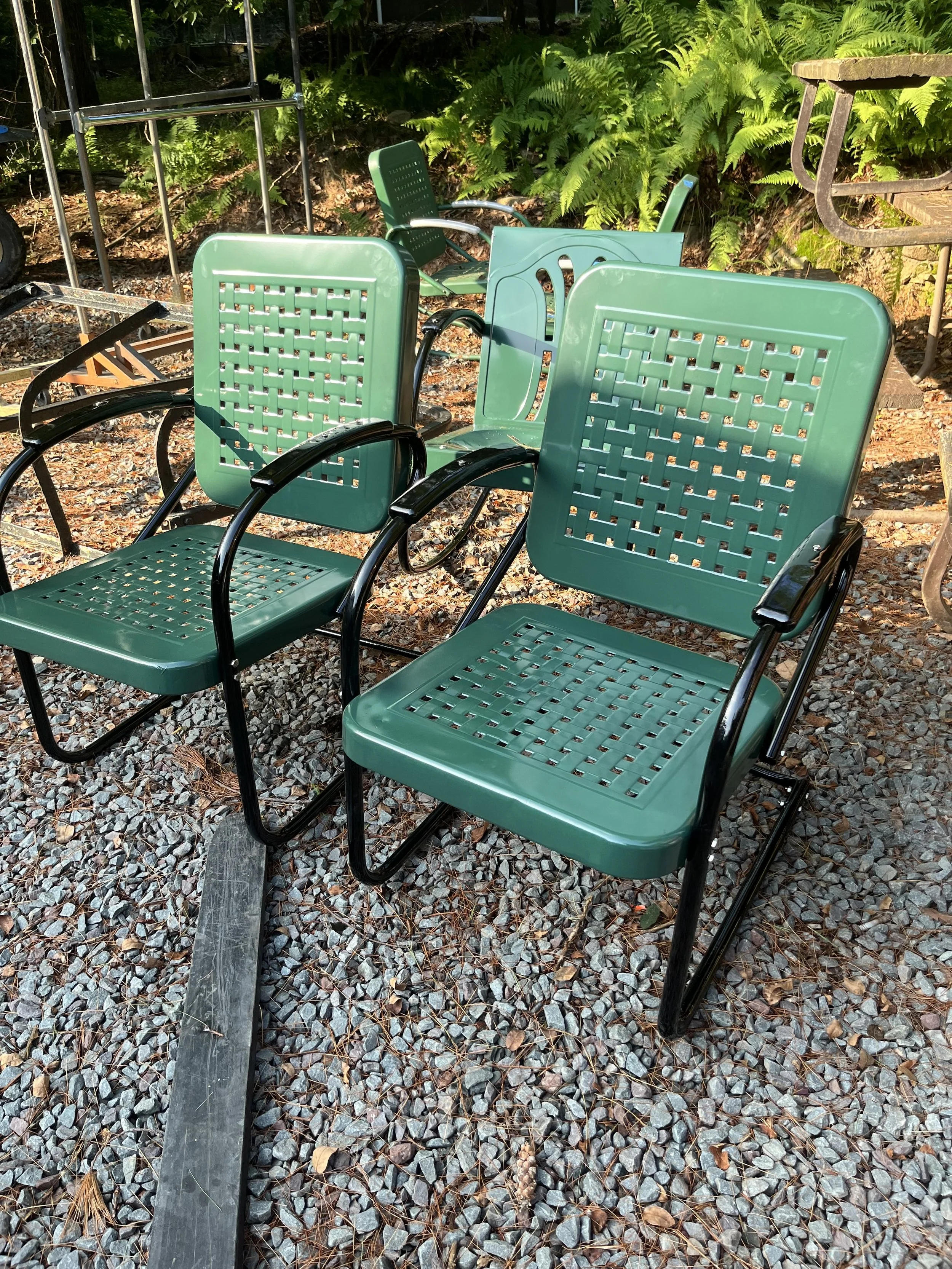 Green outdoor chairs with black metal frames on gravel ground surrounded by green foliage.