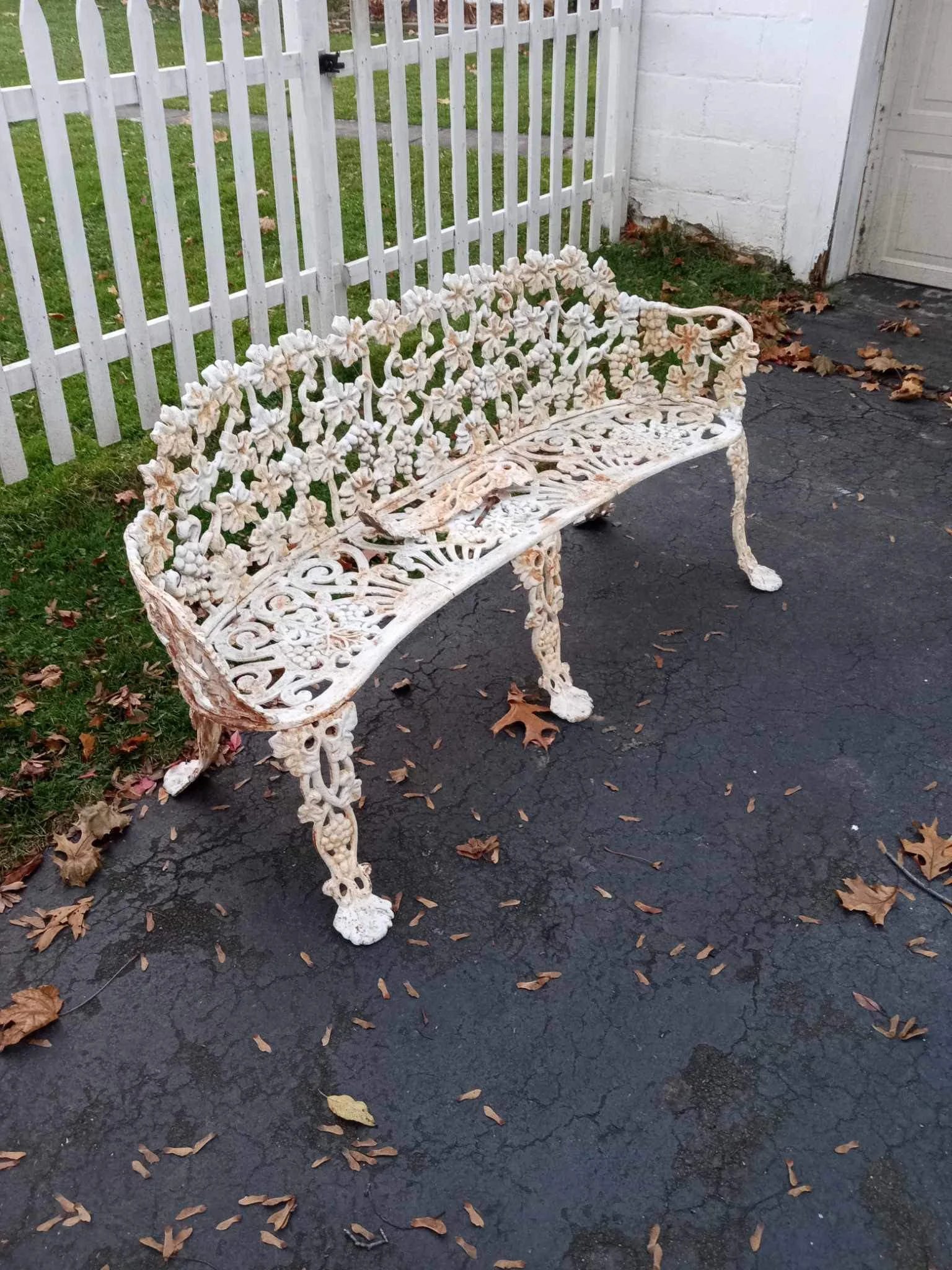 Decorated white metal garden bench with floral and vine patterns on a black asphalt surface, surrounded by fallen leaves, near a white picket fence and a white building.